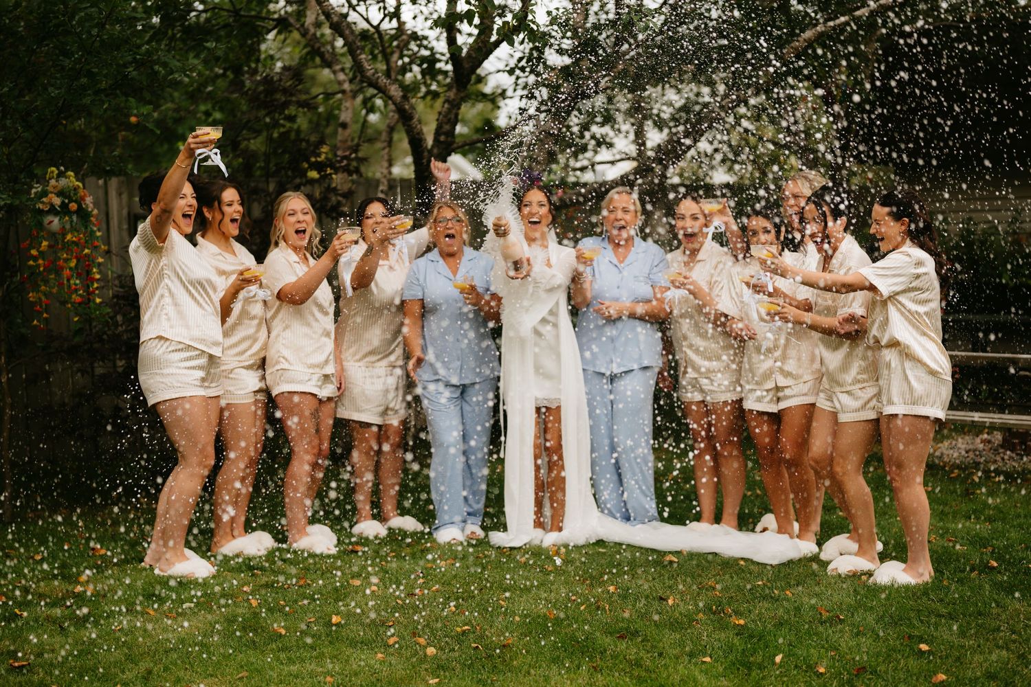 Bride in white gown celebrates with bridesmaids in matching pajamas outdoors, popping champagne in a joyful shower.