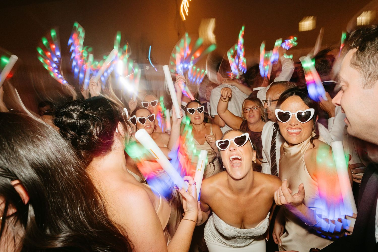 Bride and wedding guests dancing at reception party with colorful LED glow sticks and heart-shaped sunglasses.