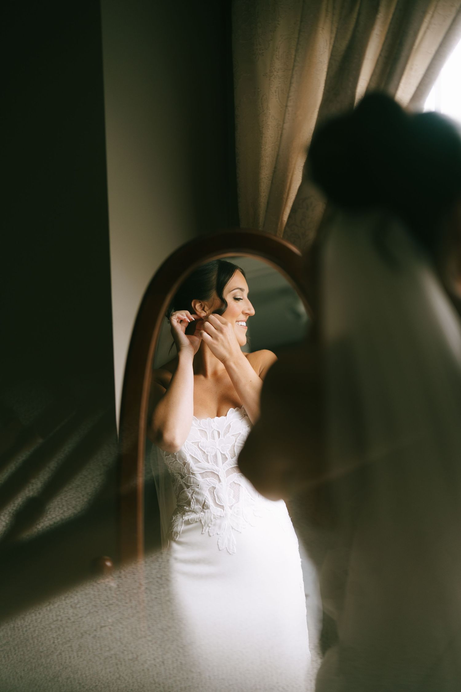 Bride in white gown adjusting earring while smiling at oval mirror, wedding veil visible in foreground.