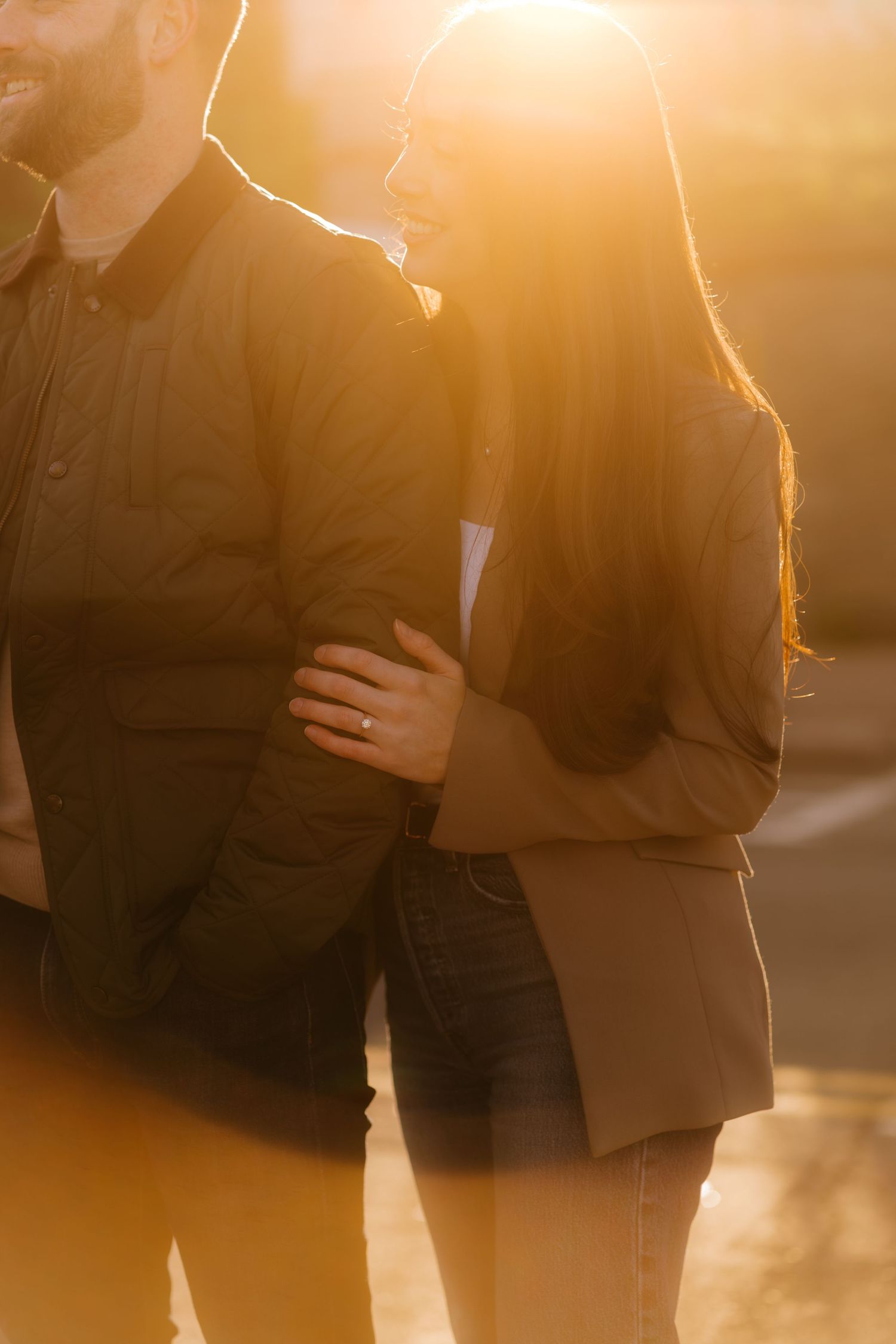 Couple embracing at golden hour sunset, woman wearing engagement ring, warm backlit glow highlighting romantic moment outdoors.