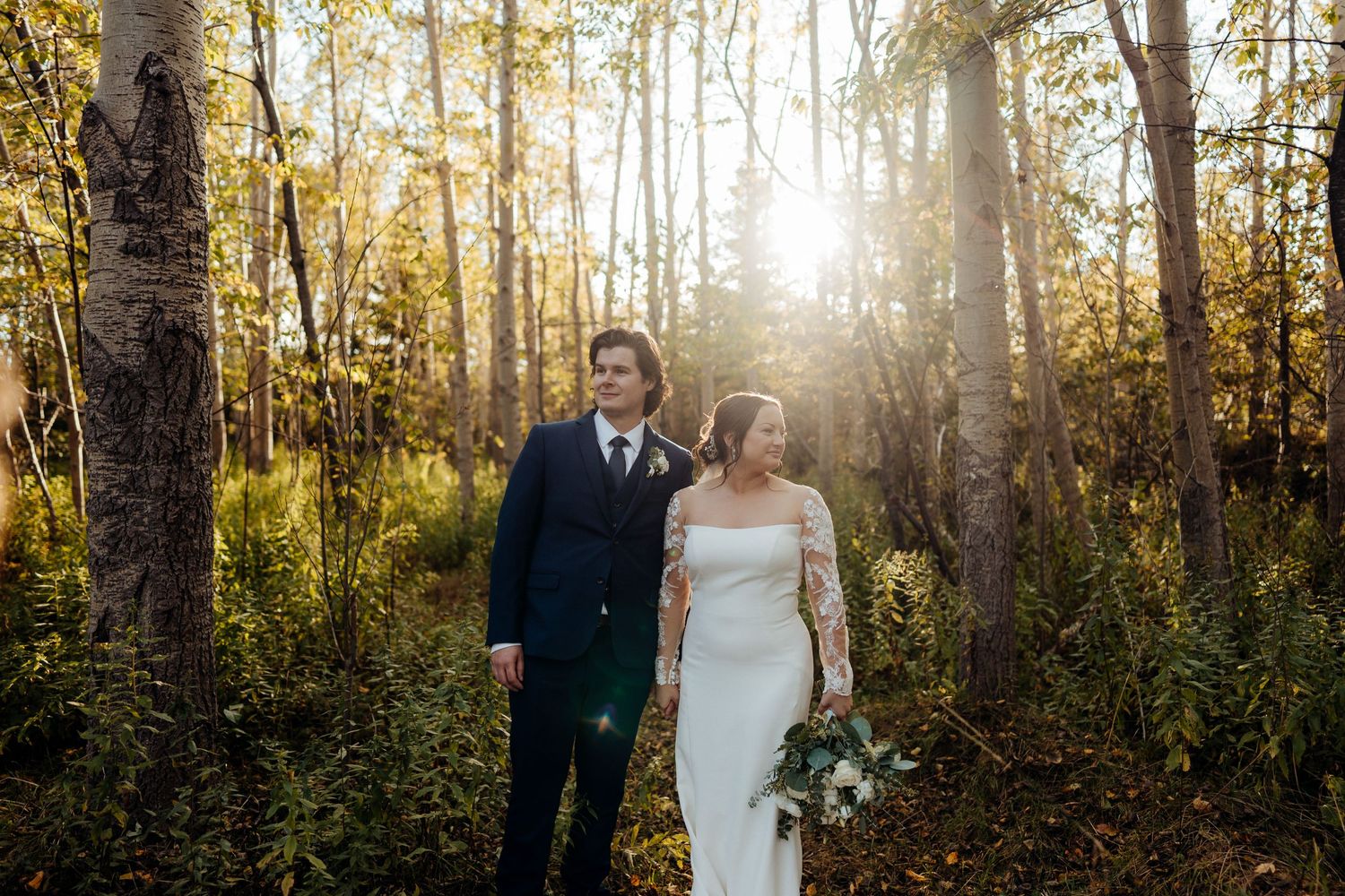 A wedding couple embraces in a sunlit autumn forest with golden light streaming through the trees.