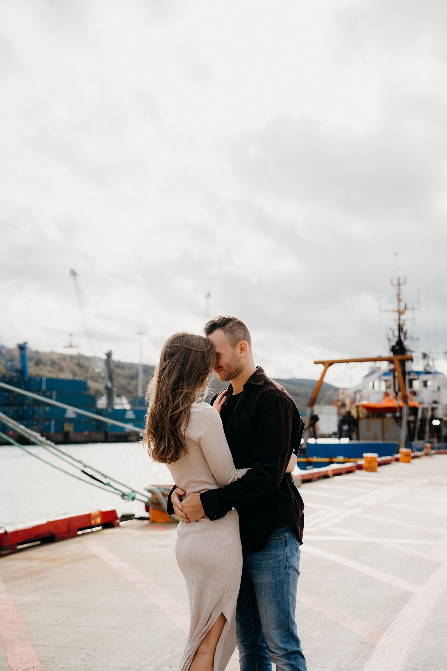 Couple in formal and casual wear sharing intimate moment at industrial port setting.