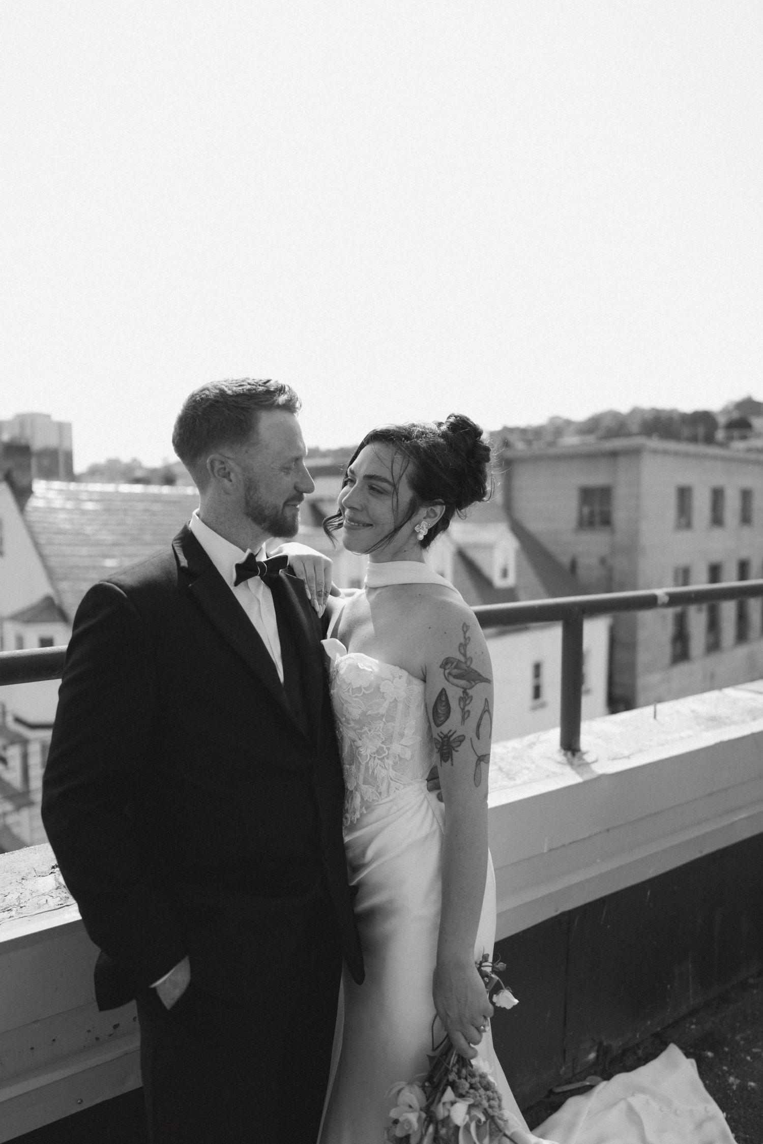 Black and white photo of a bride and groom embracing on a rooftop, smiling at each other in formal wedding attire.
