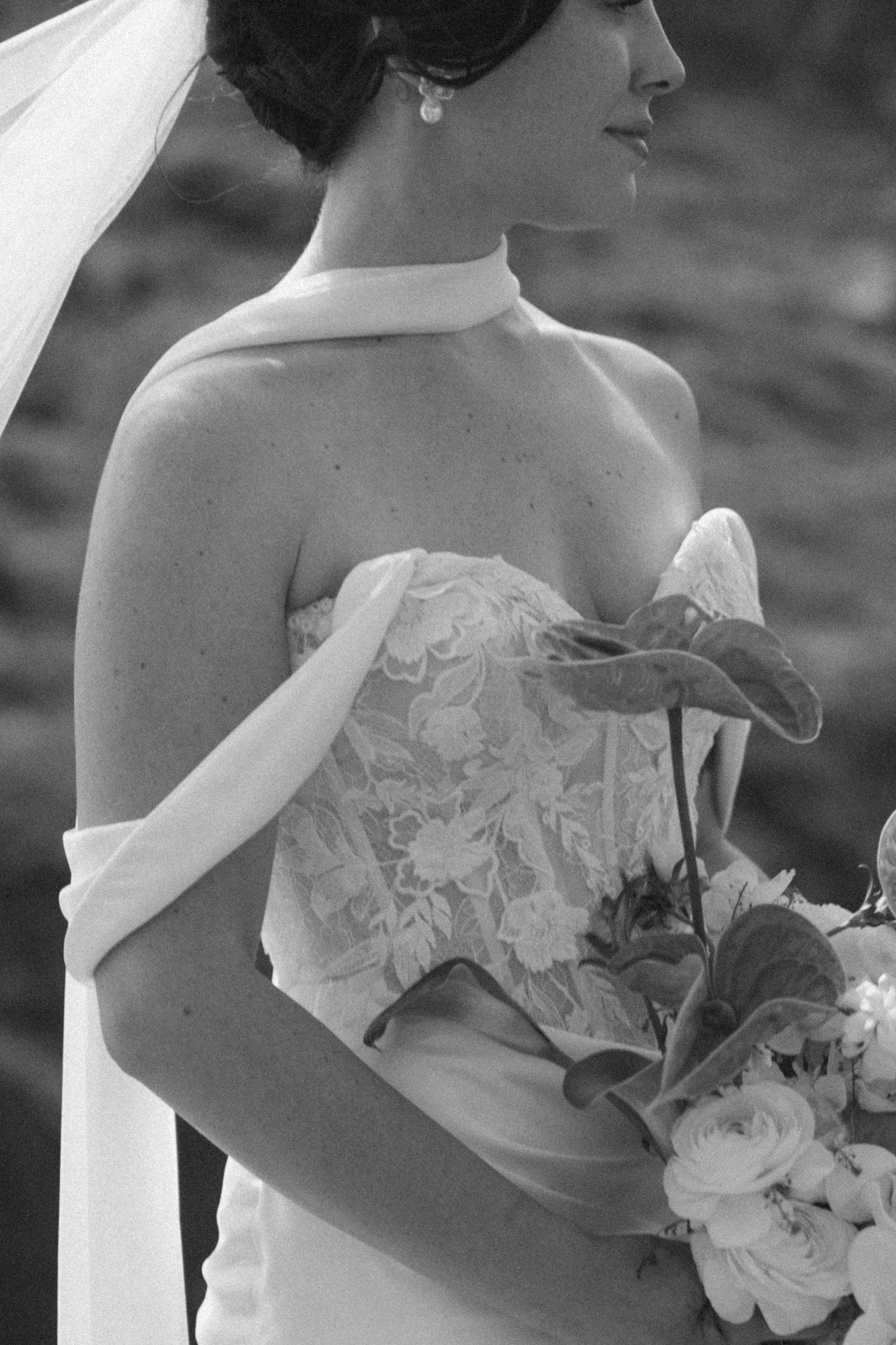Black and white photo of a bride in a lace strapless gown holding a floral bouquet, wearing a veil and pearl earring.