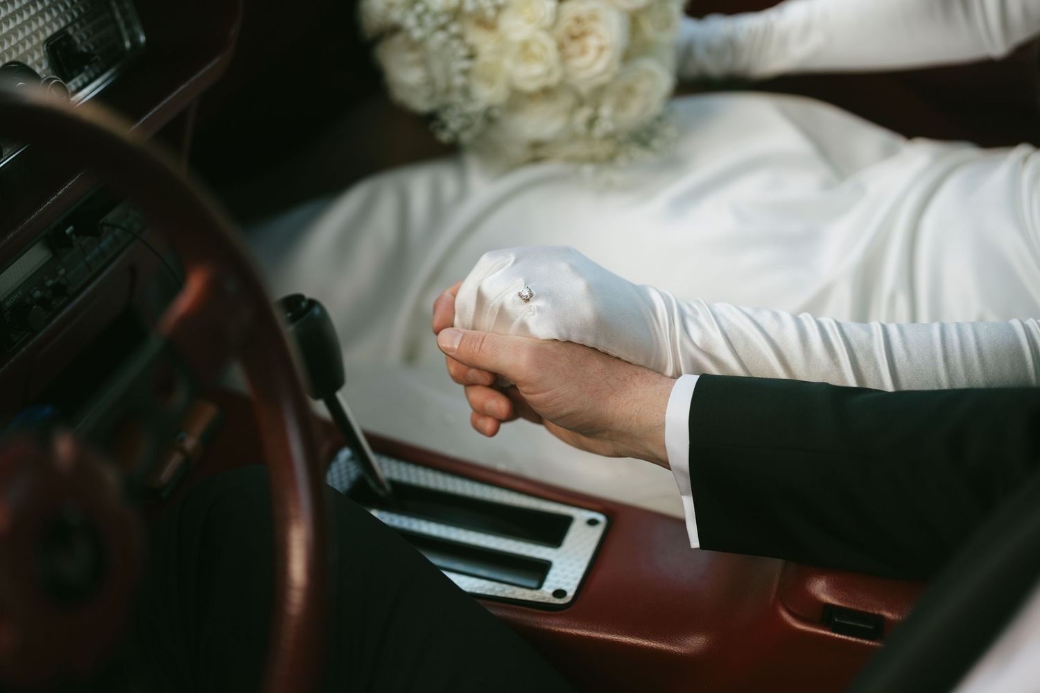 Bride in white gown holding bouquet beside groom shifting gears in vintage car interior on wedding day.