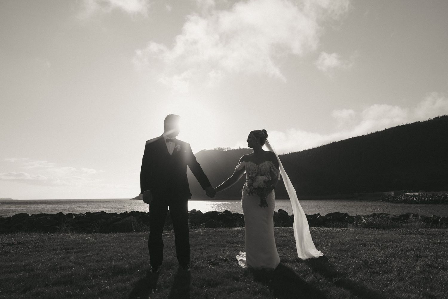 Black and white photo of a bride and groom holding hands, silhouetted against a coastal sunset landscape.