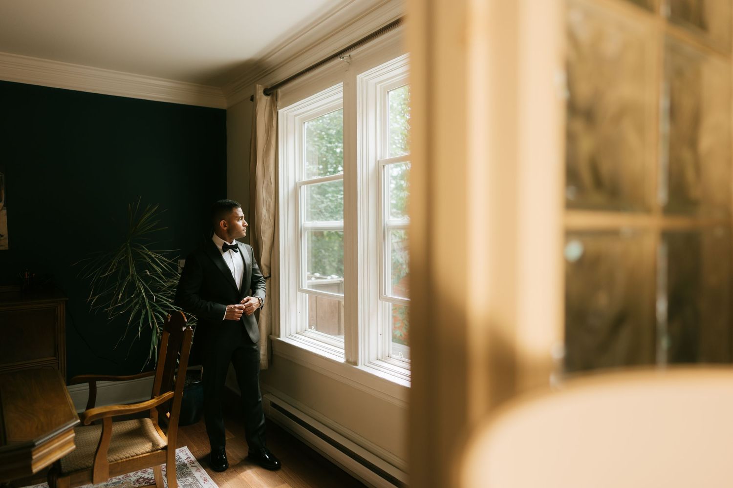 Groom in black tuxedo and bow tie gazing out window in elegant room before wedding ceremony.