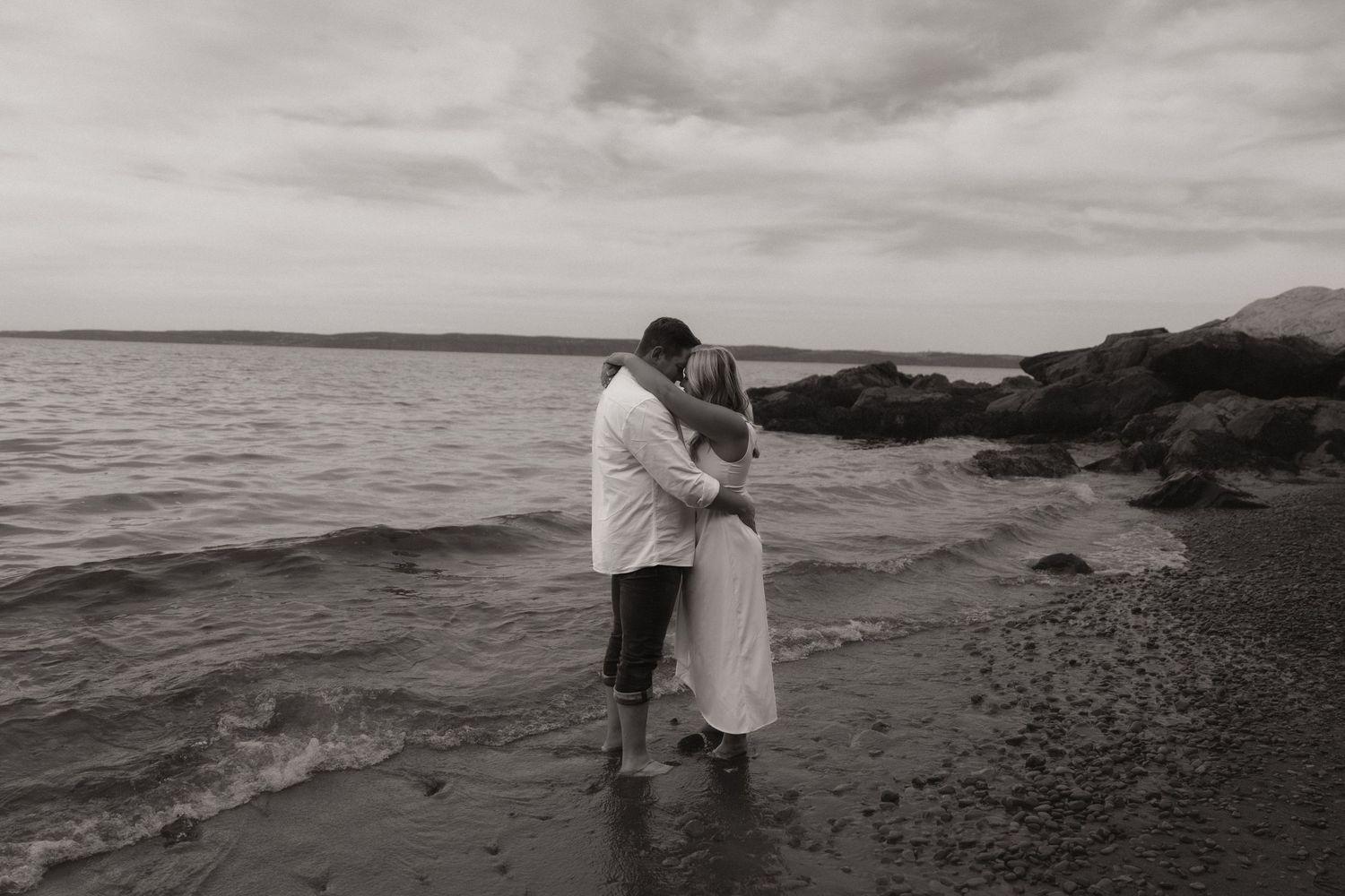 Black and white photo of a couple walking along a rocky beach shoreline with waves crashing nearby.