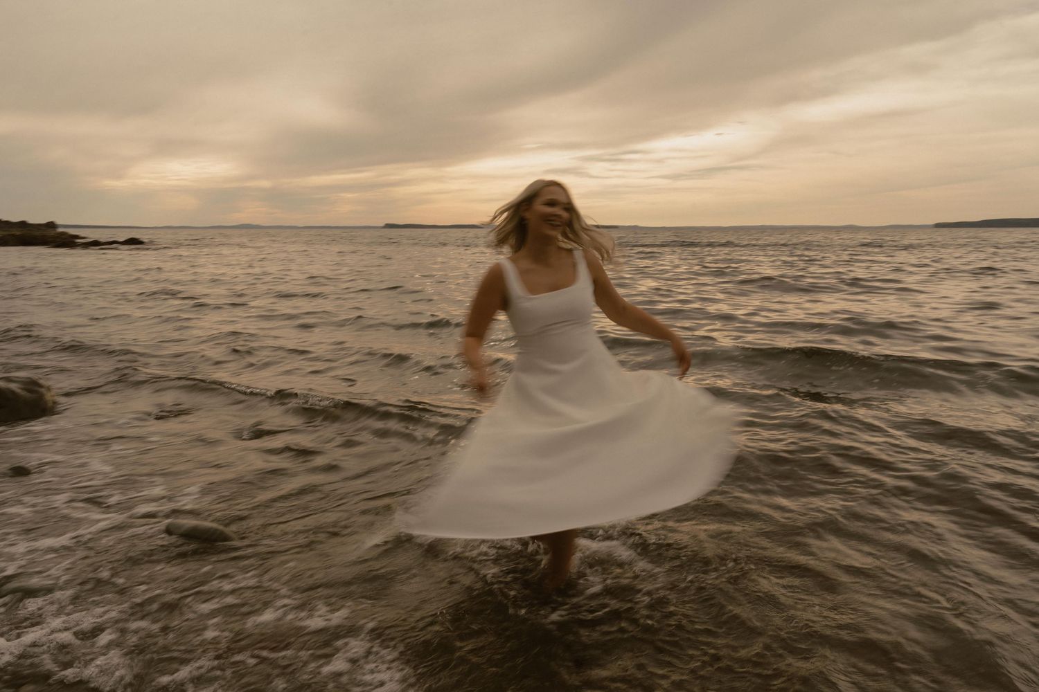 Bride in flowing white dress walks through shallow ocean water at sunset creating a dramatic silhouette.