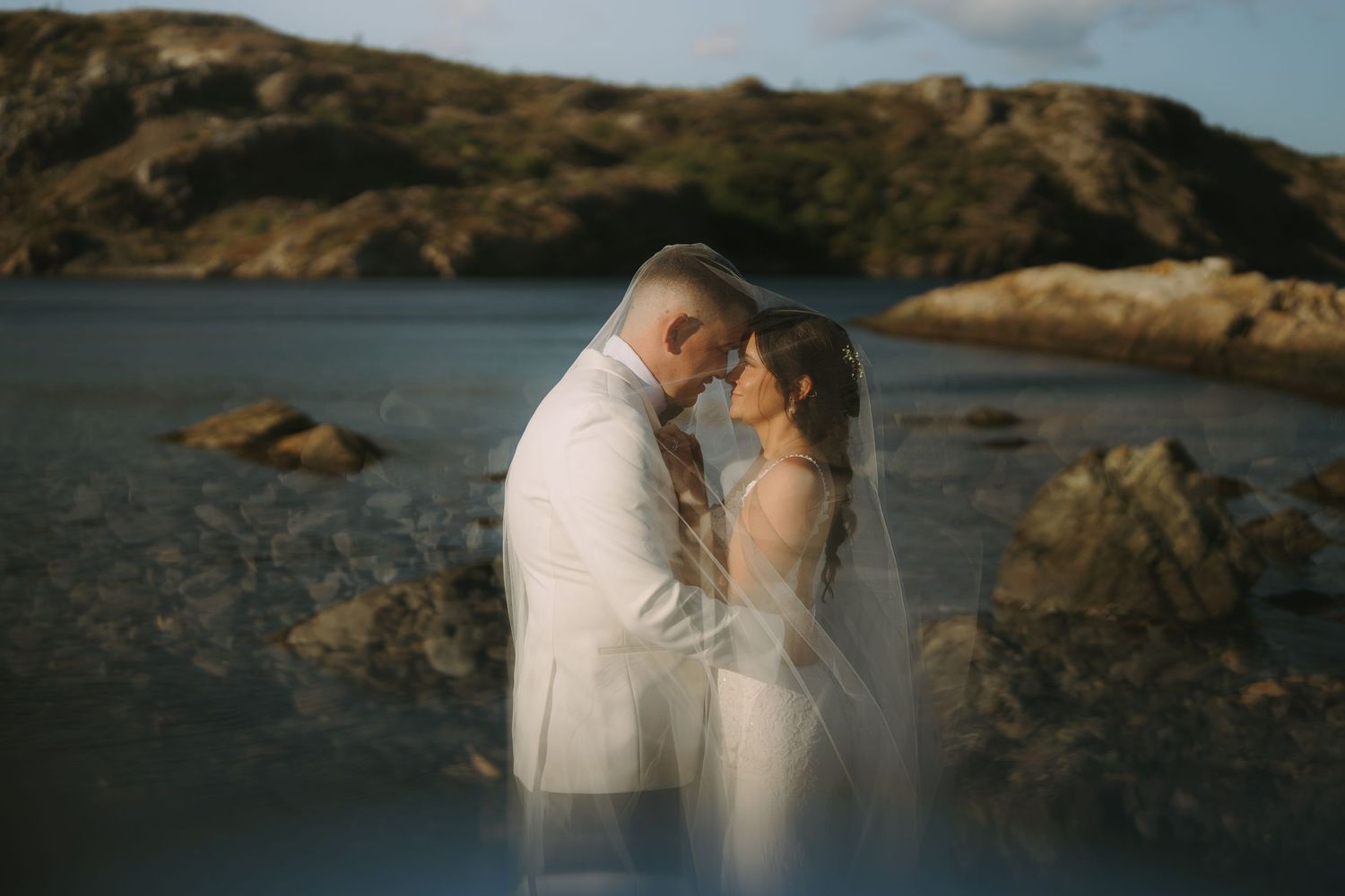 A romantic couple embraces on a rocky coastal shore at sunset with mountains in the background.