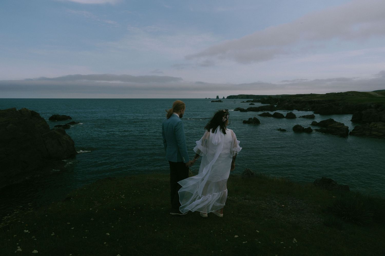 A couple in wedding attire stands along a rocky coastline at dusk with dramatic cloudy skies in the background.