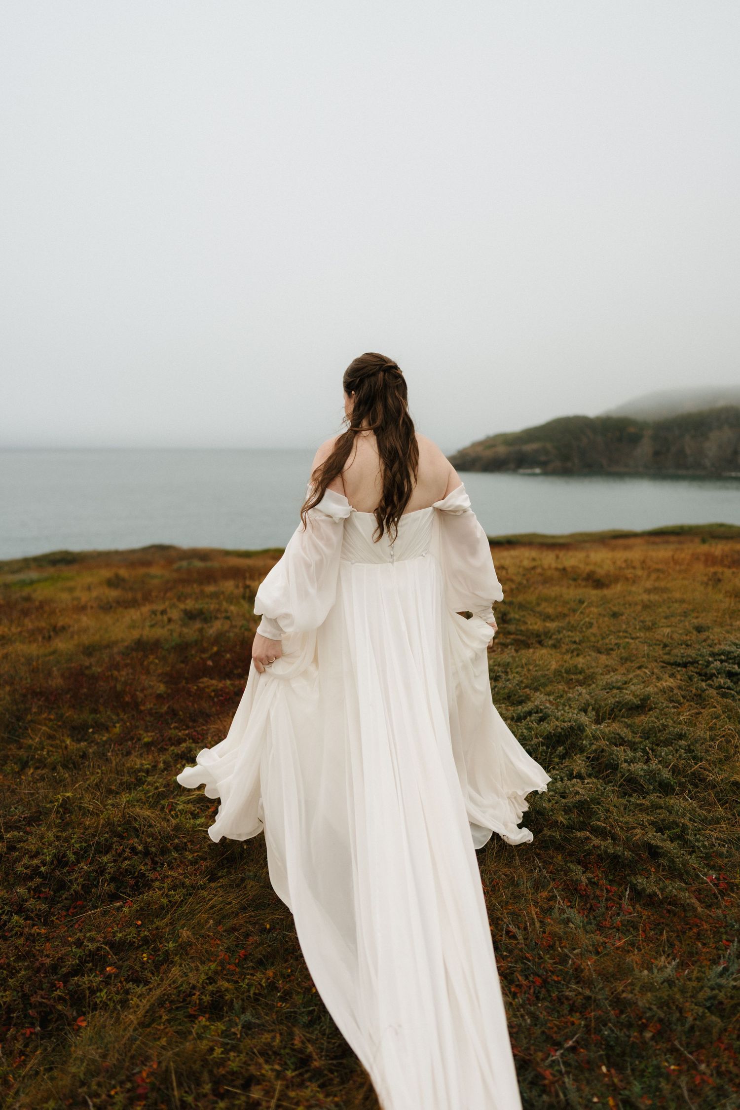 A flowing white wedding dress billows in the wind as someone stands on a misty coastal cliff overlooking the ocean.