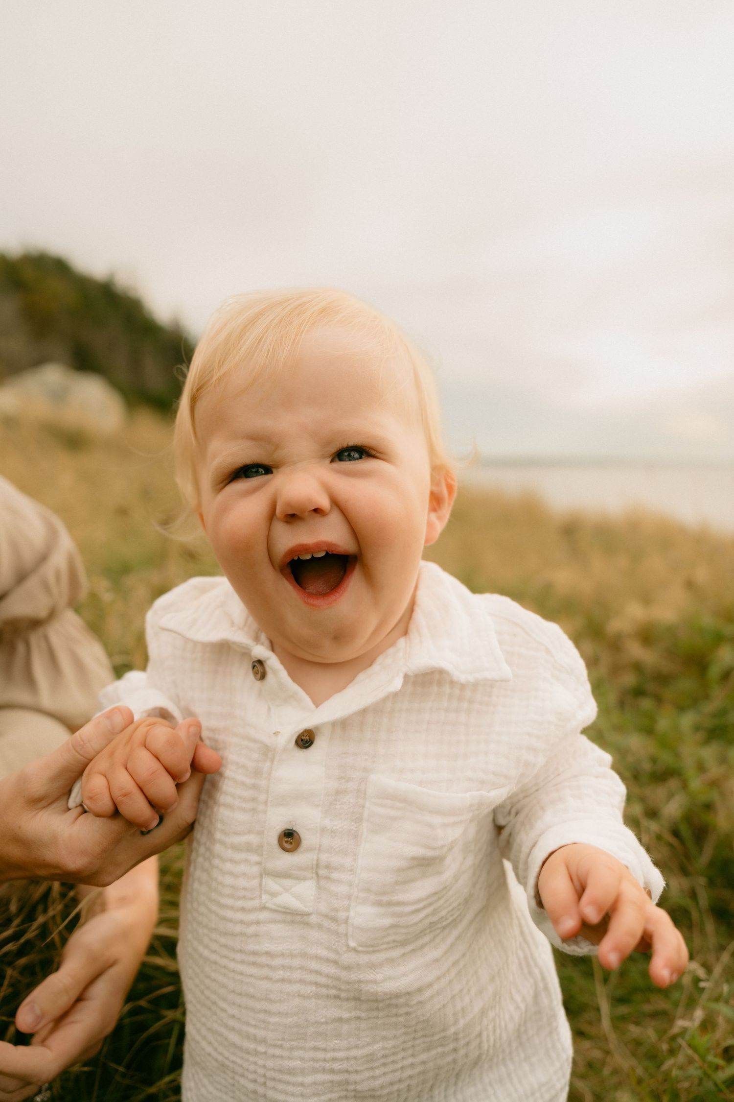 A joyful baby in a white linen shirt laughs while standing outdoors against a coastal backdrop.