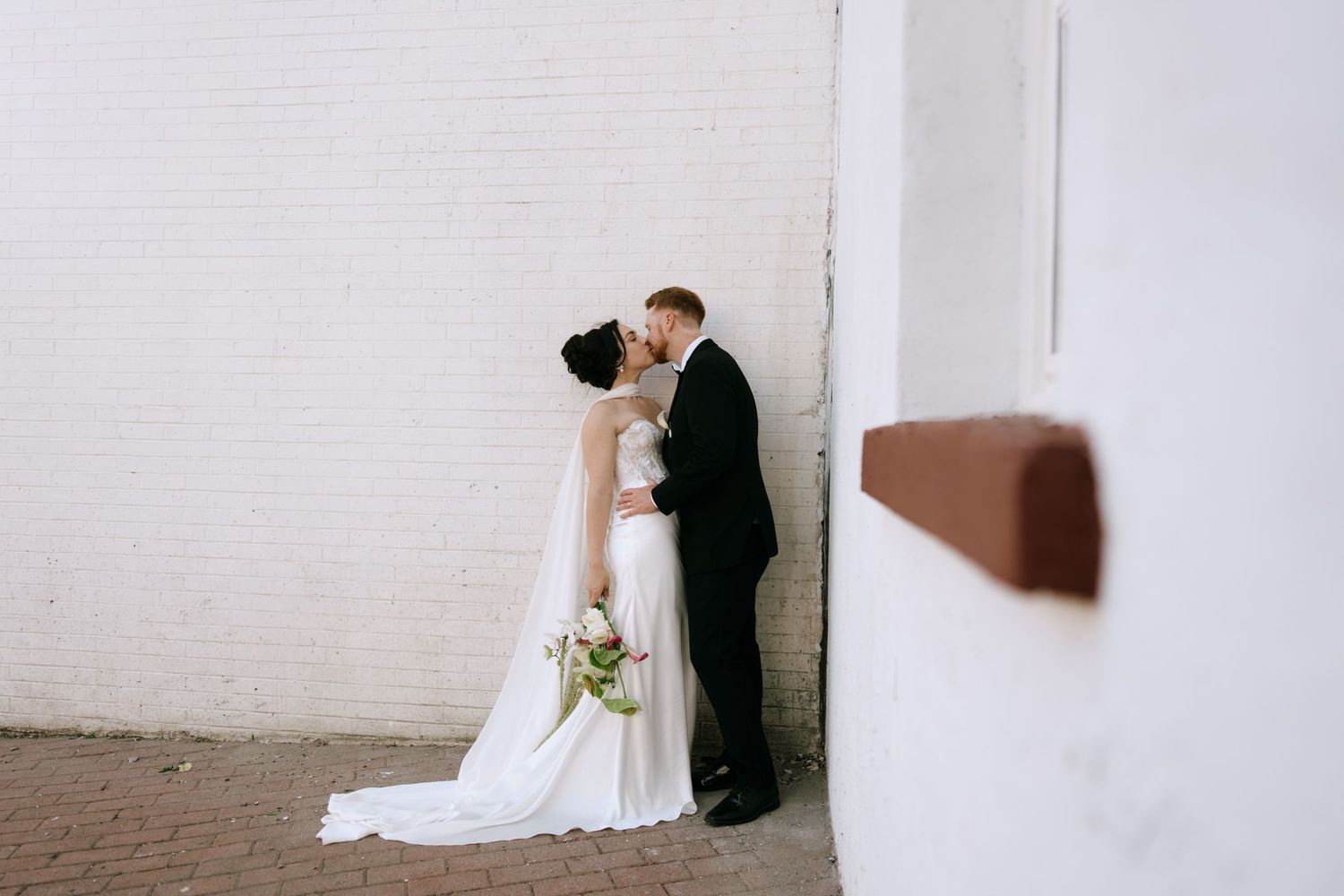 Bride and groom share a kiss against a white brick wall, bride holding a green and white bouquet.