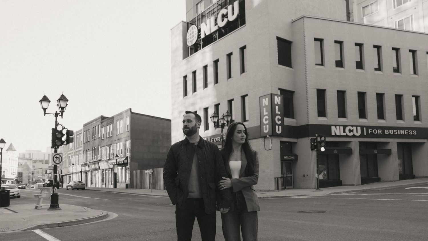 A couple stands on an urban street corner near an NLCU bank building in a black and white photo.