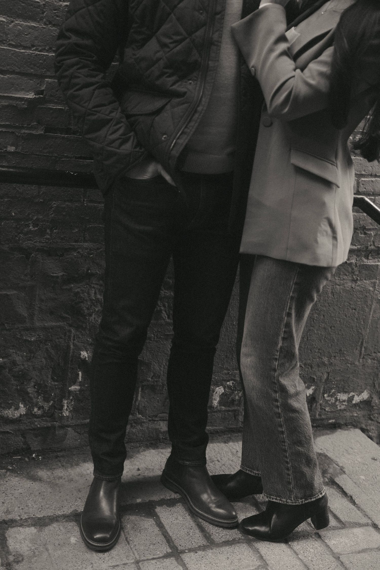 Black and white photo of two people standing against a brick wall, wearing boots, jeans, and jackets.