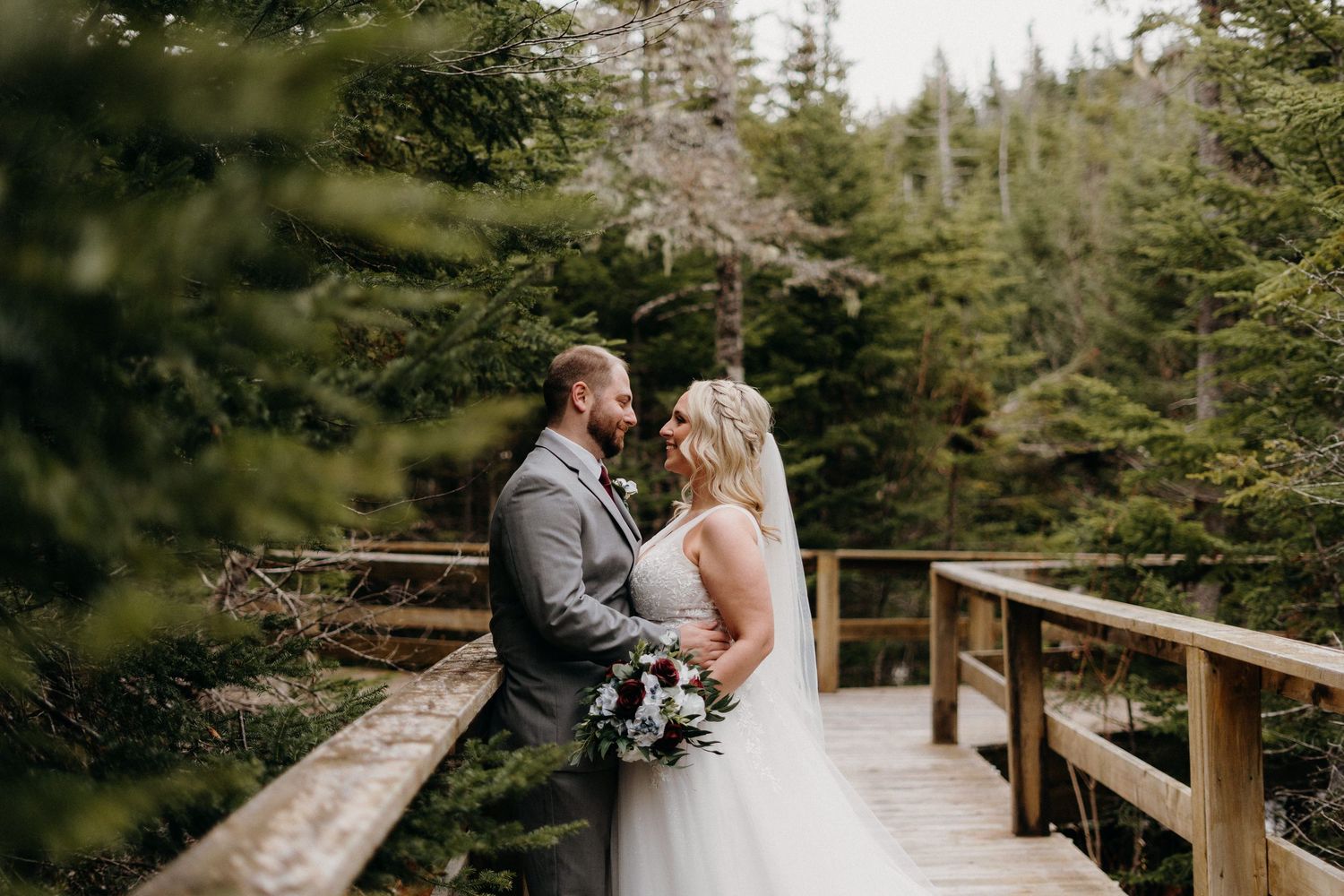 A couple shares an intimate moment on a wooden bridge surrounded by evergreen trees.