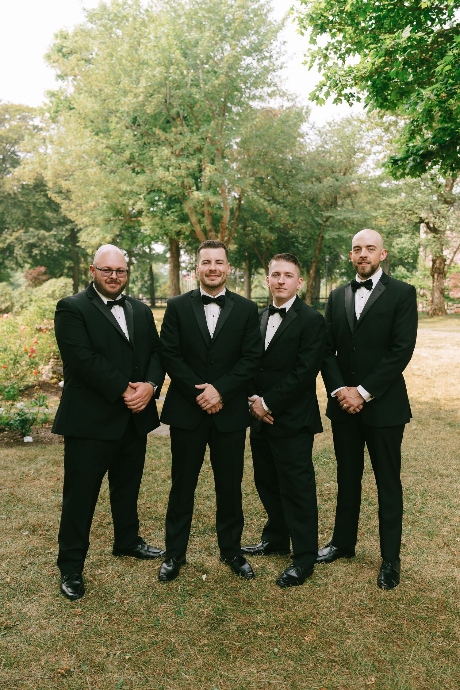 Row of groomsmen in black tuxedos standing together on grass for wedding photos.