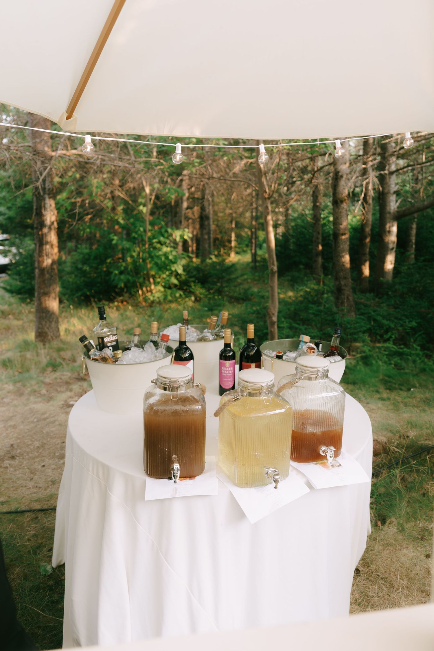 Beverage dispensers with various drinks set up on white tables under a tent in a wooded outdoor setting.