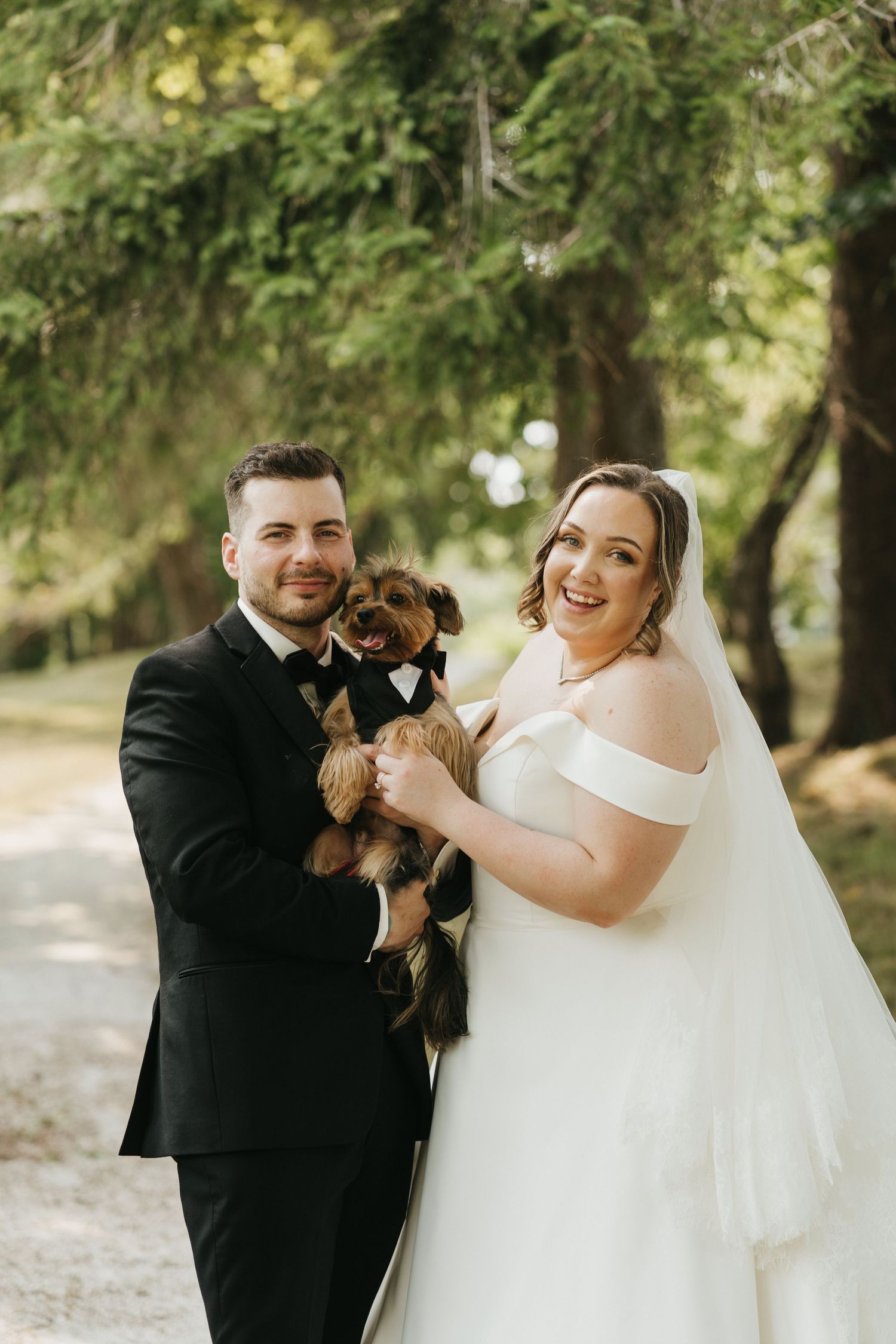 Wedding couple laughs while holding their small dog during outdoor photo session.