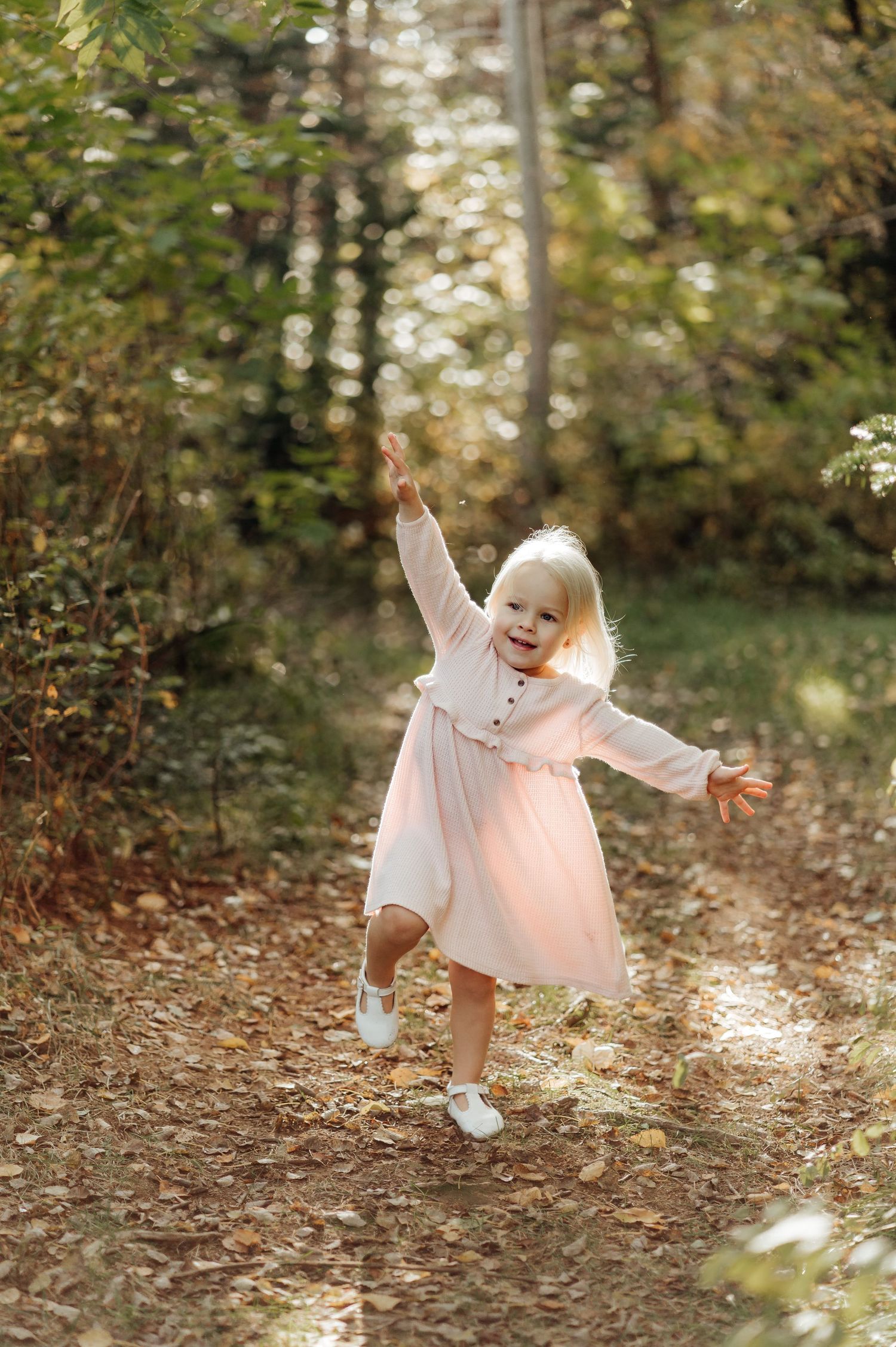 A playful child in a pink dress dances through autumn leaves in a forest setting.