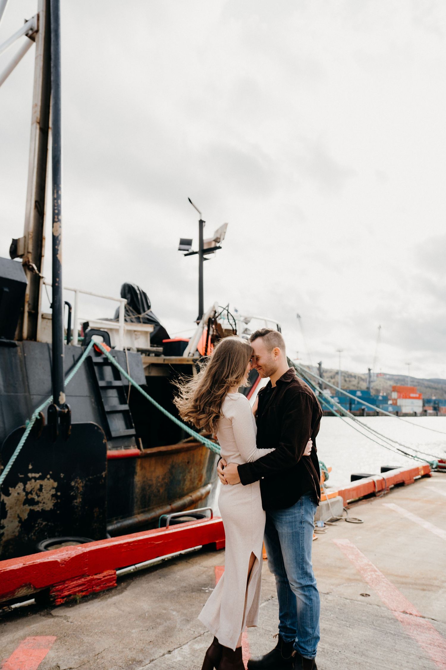 A couple stands together on an industrial dock with ships and harbor equipment in view.