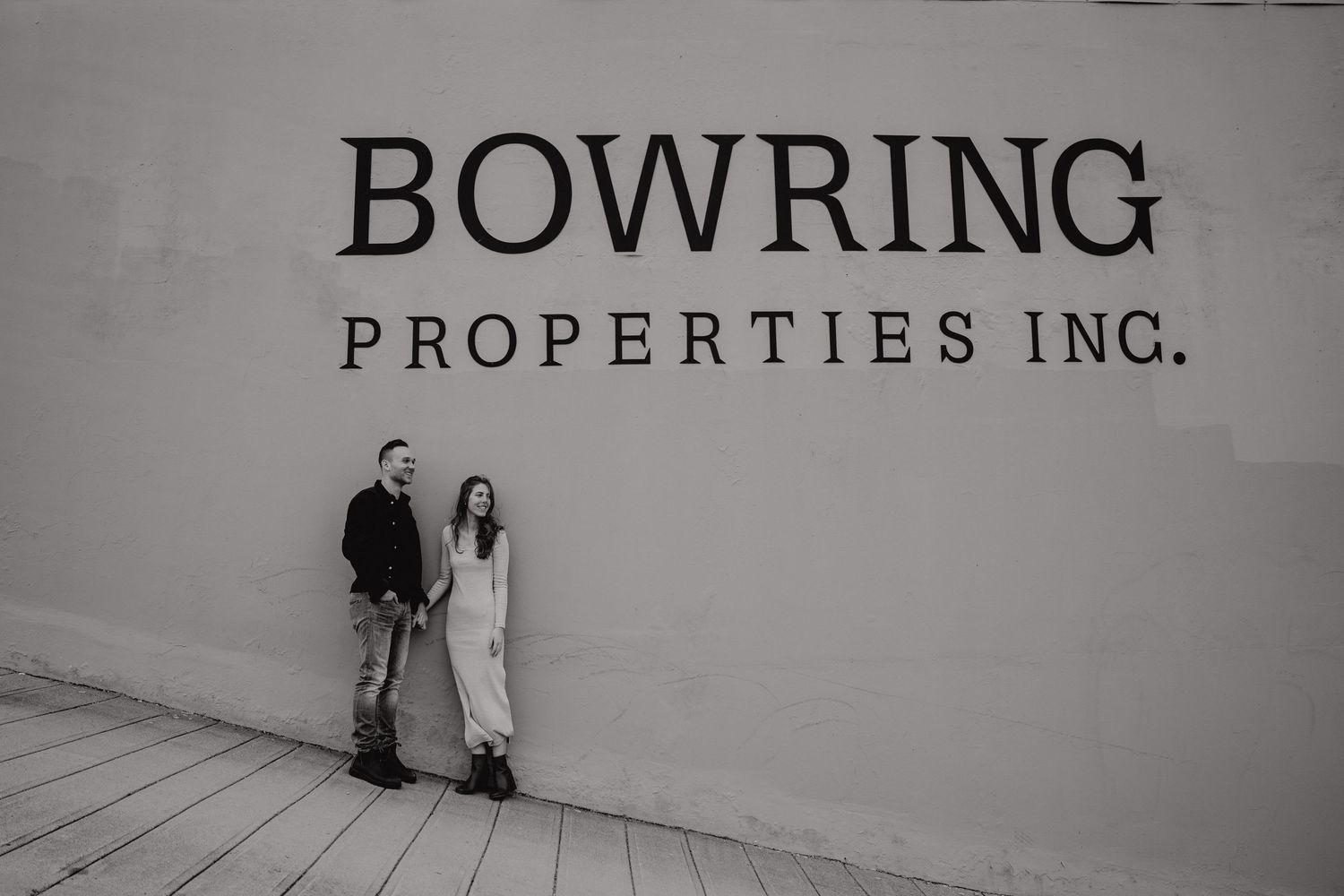 Two people photographed in black and white against an industrial wall with company branding.