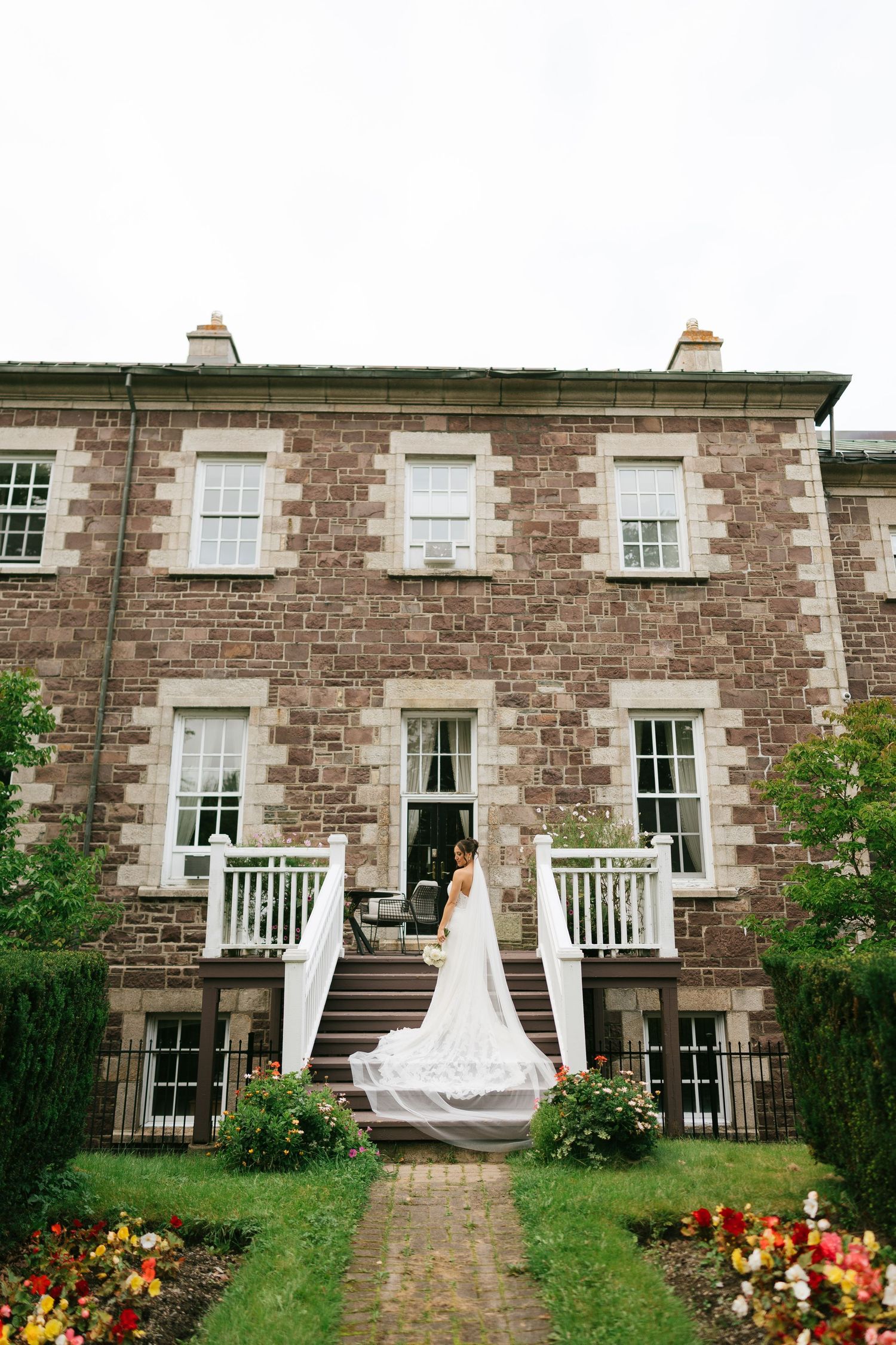 Bride in white gown with long veil stands on steps of elegant stone manor house surrounded by lush gardens.