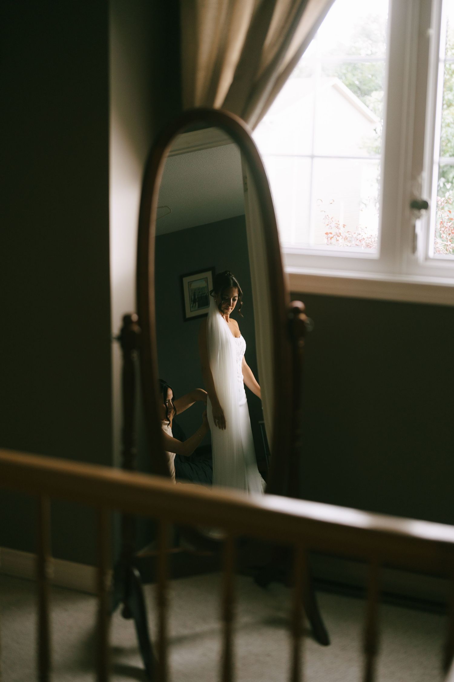 Bride in white wedding dress and veil reflected in an oval wooden mirror, standing by a window in a softly lit room.