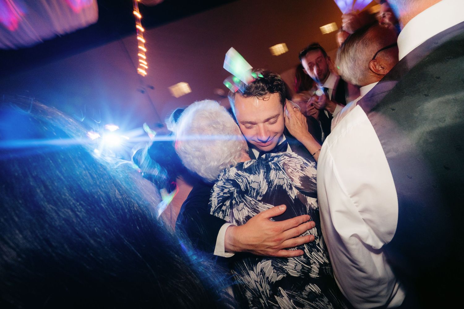 Two guests share a warm embrace on the dance floor at a lively wedding reception with colorful lights.