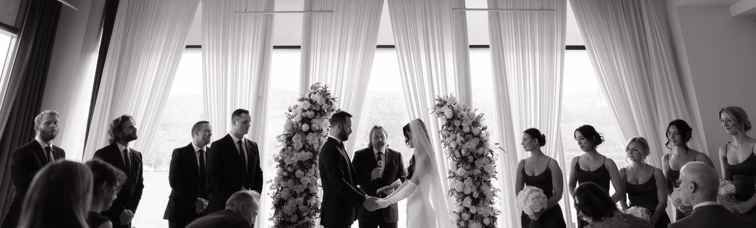 Black and white wedding ceremony photo with couple at floral arch, officiant, groomsmen, and bridesmaids in elegant venue.