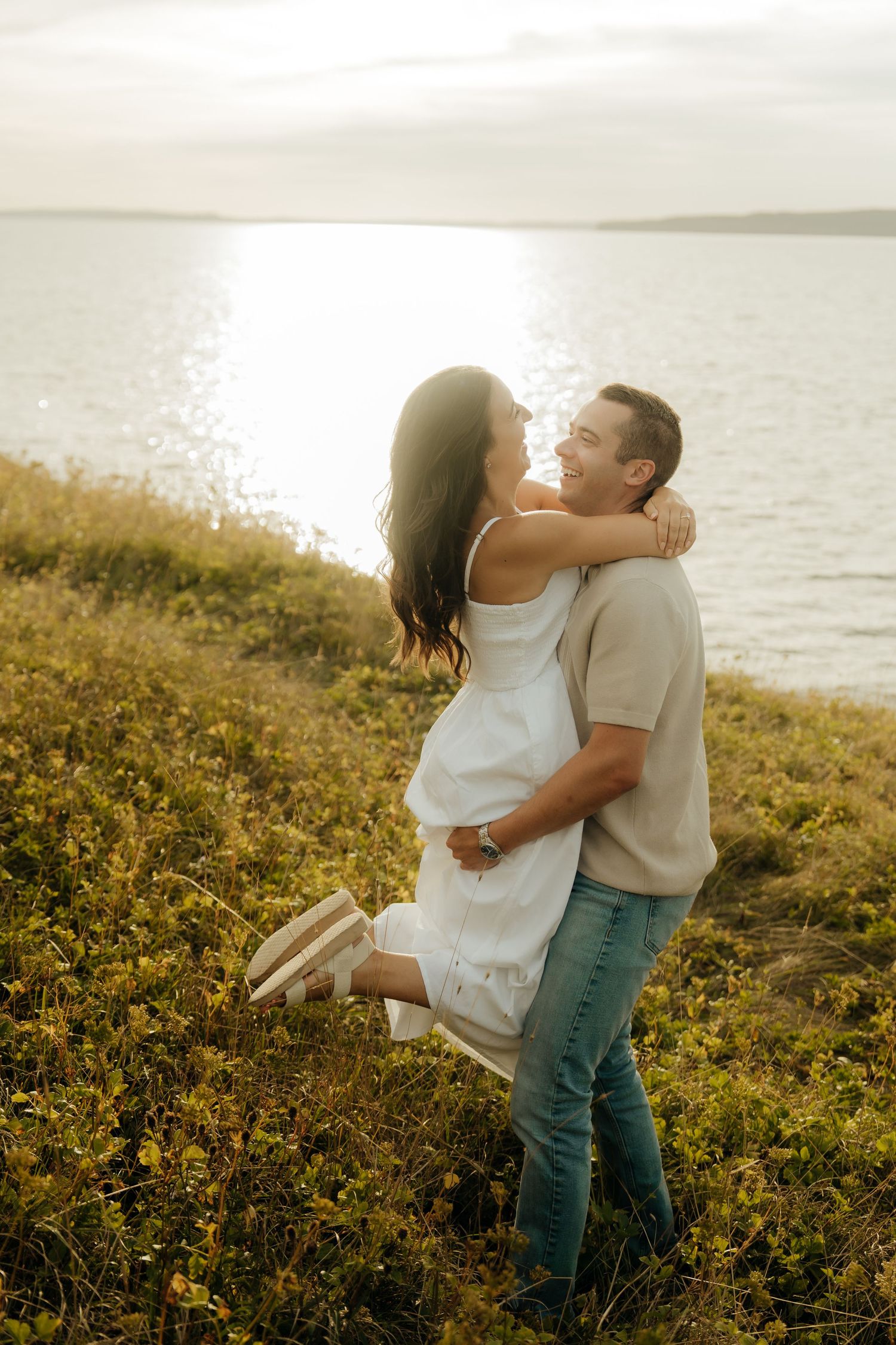 A couple embraces lovingly in a golden meadow by the ocean during sunset, creating a romantic silhouette.