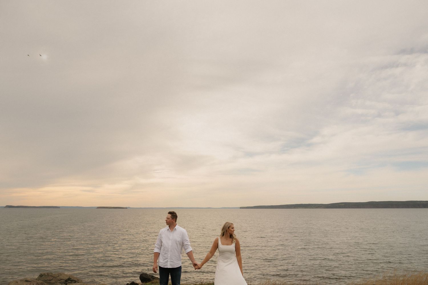 Couple walking hand in hand along a serene beach at sunset with cloudy skies and calm waters.