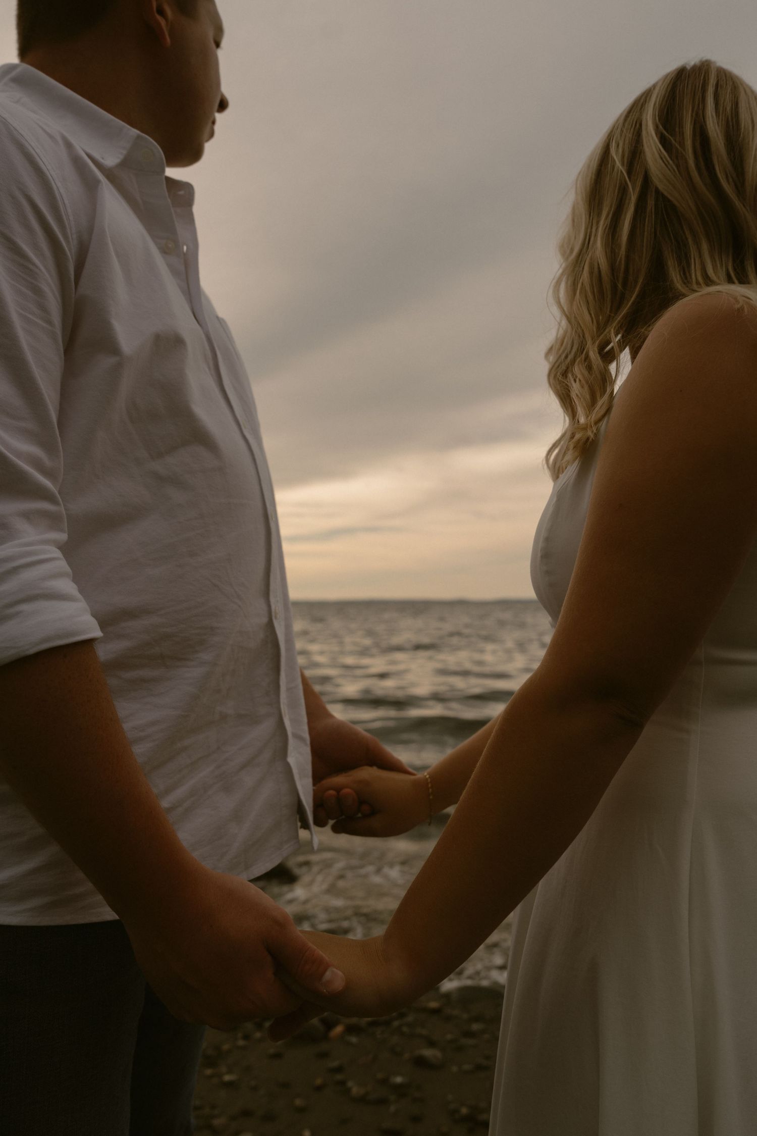 A romantic seaside moment captured at sunset as a couple in white clothing holds hands by the ocean.