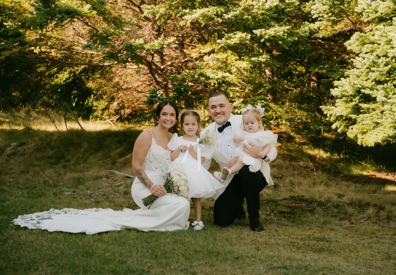 Family group lounges together on grass in a scenic wooded setting during golden hour sunlight.
