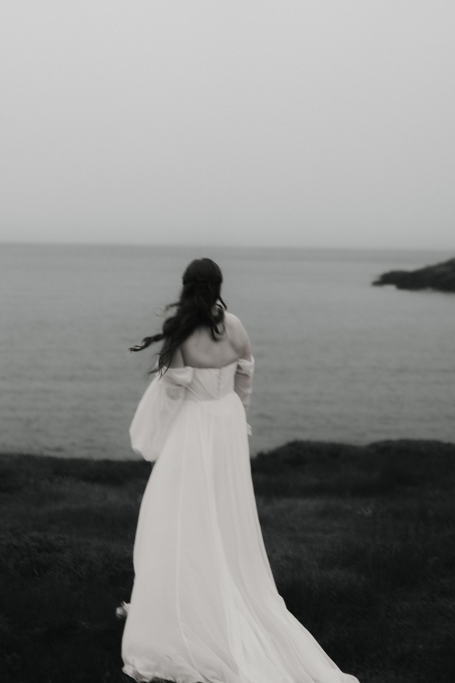 A figure in a flowing white dress stands against a moody coastal landscape in black and white.