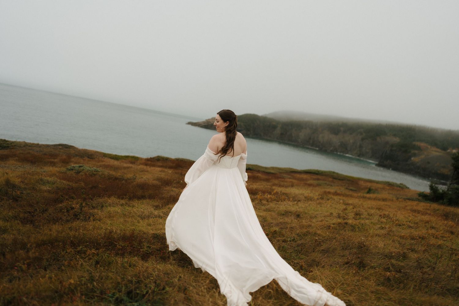 A flowing white wedding dress billows in the wind on a dramatic coastal clifftop overlooking the misty ocean.