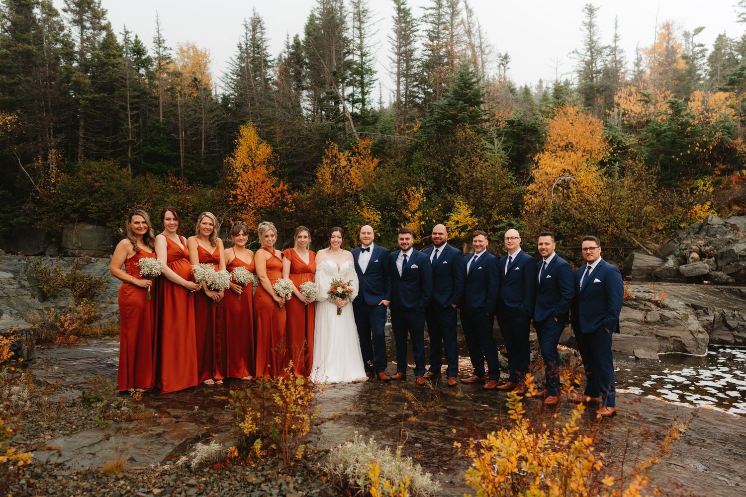 Wedding party stands together in front of vibrant autumn foliage and rocky landscape during fall outdoor ceremony.