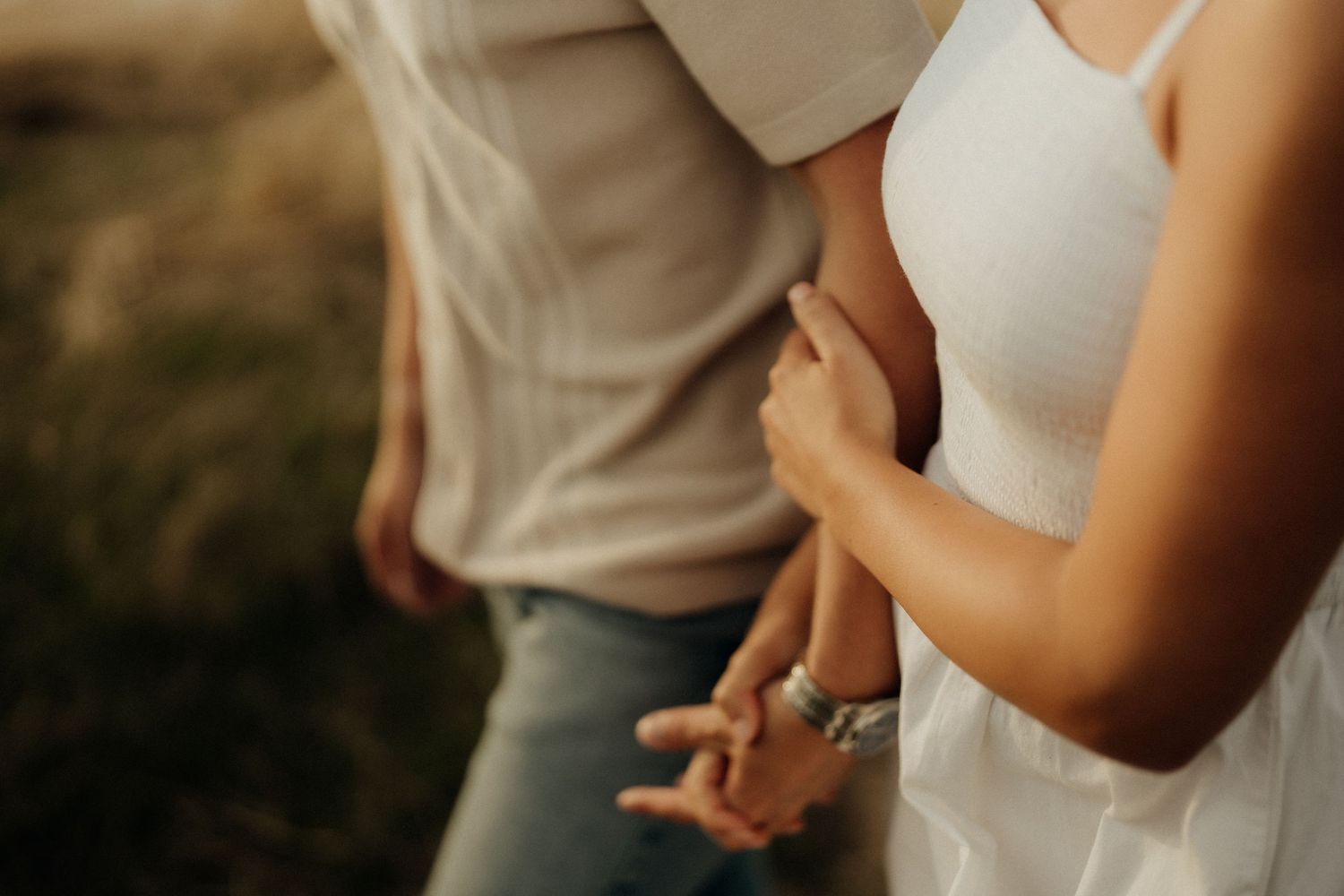A couple holds hands tenderly in a romantic outdoor setting with warm lighting and neutral-toned clothing.