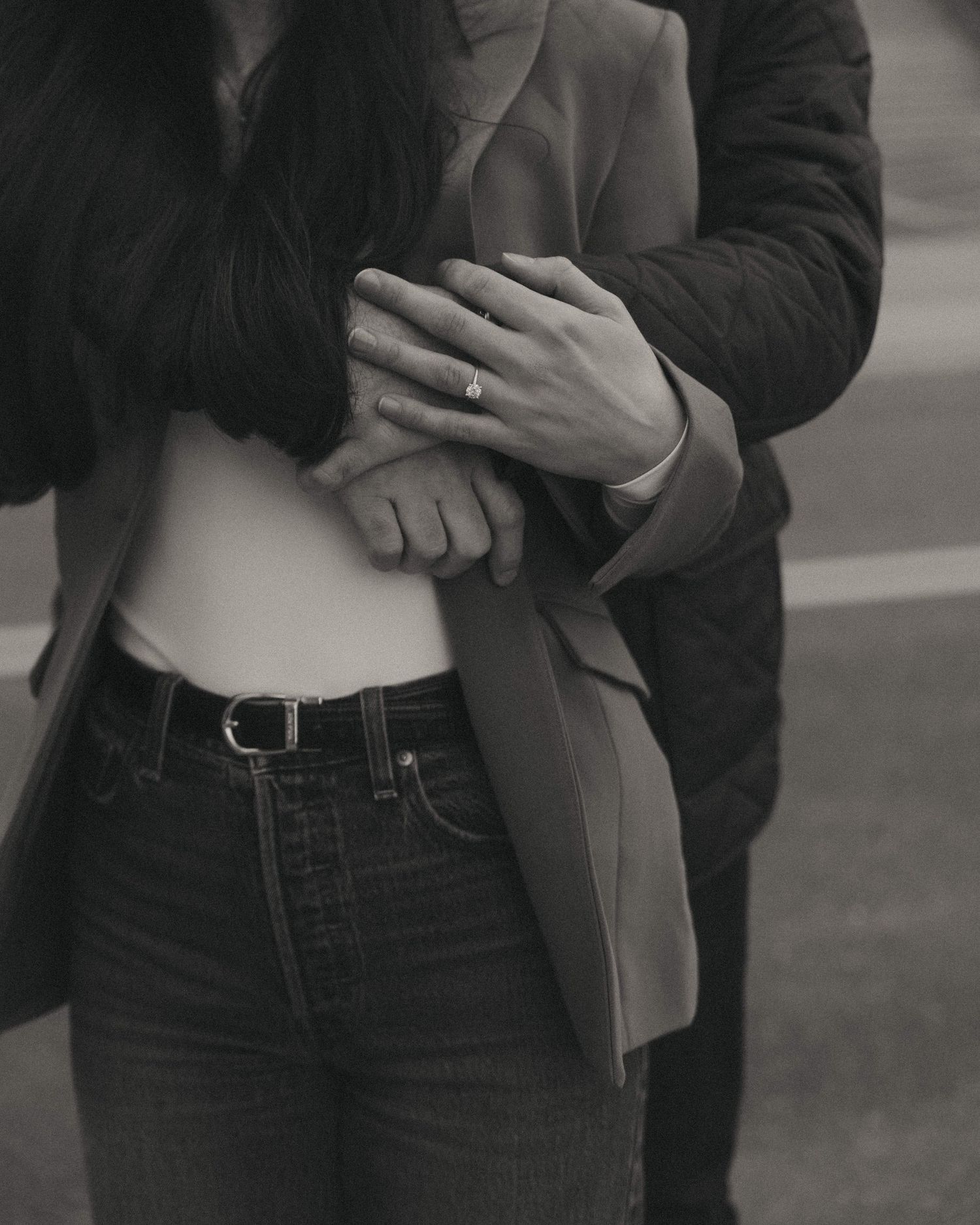 Black and white photo of a couple holding hands tenderly, woman wearing jeans and blazer with engagement ring visible.