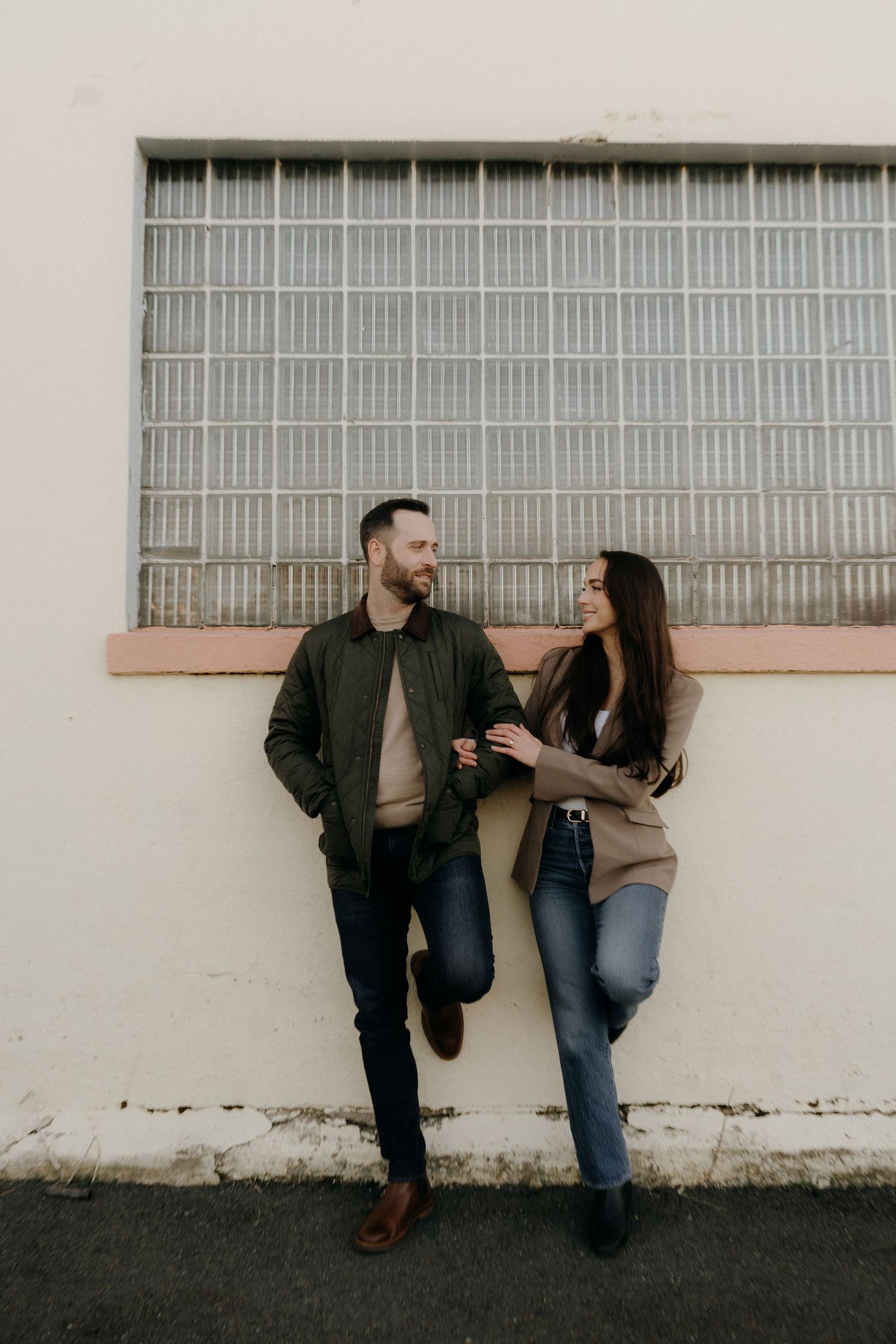 A couple leans against a white wall, smiling at each other, dressed in casual autumn outfits.