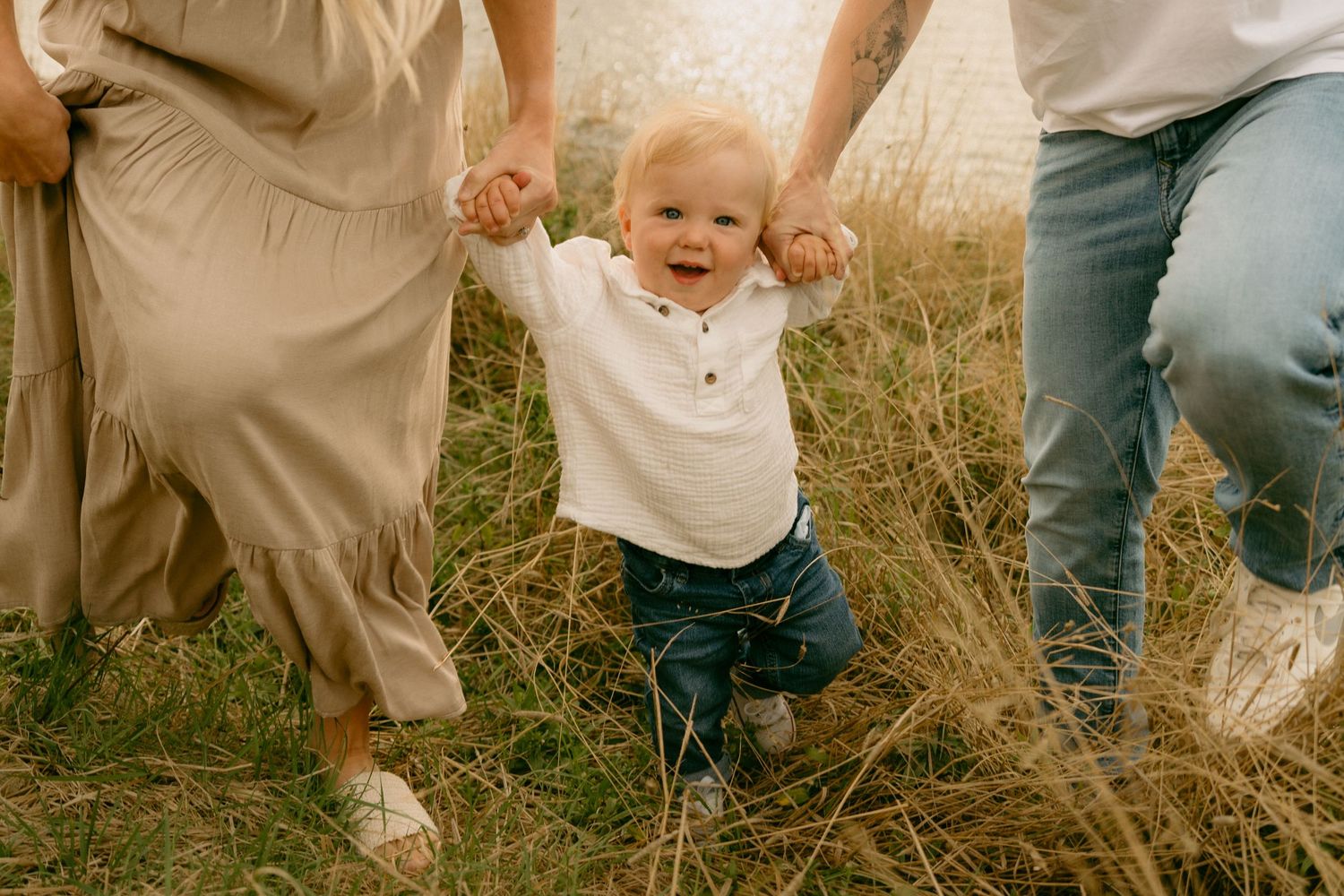 A small child in a white shirt takes joyful steps while holding hands with adults in a grassy field.