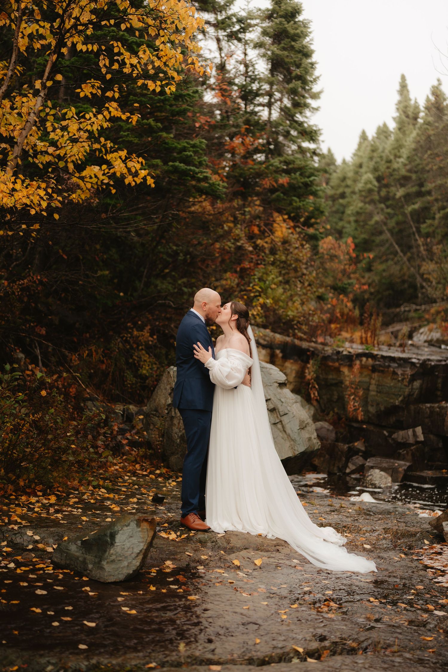 A couple shares a romantic moment on a rocky riverbank surrounded by vibrant autumn foliage in a forest setting.