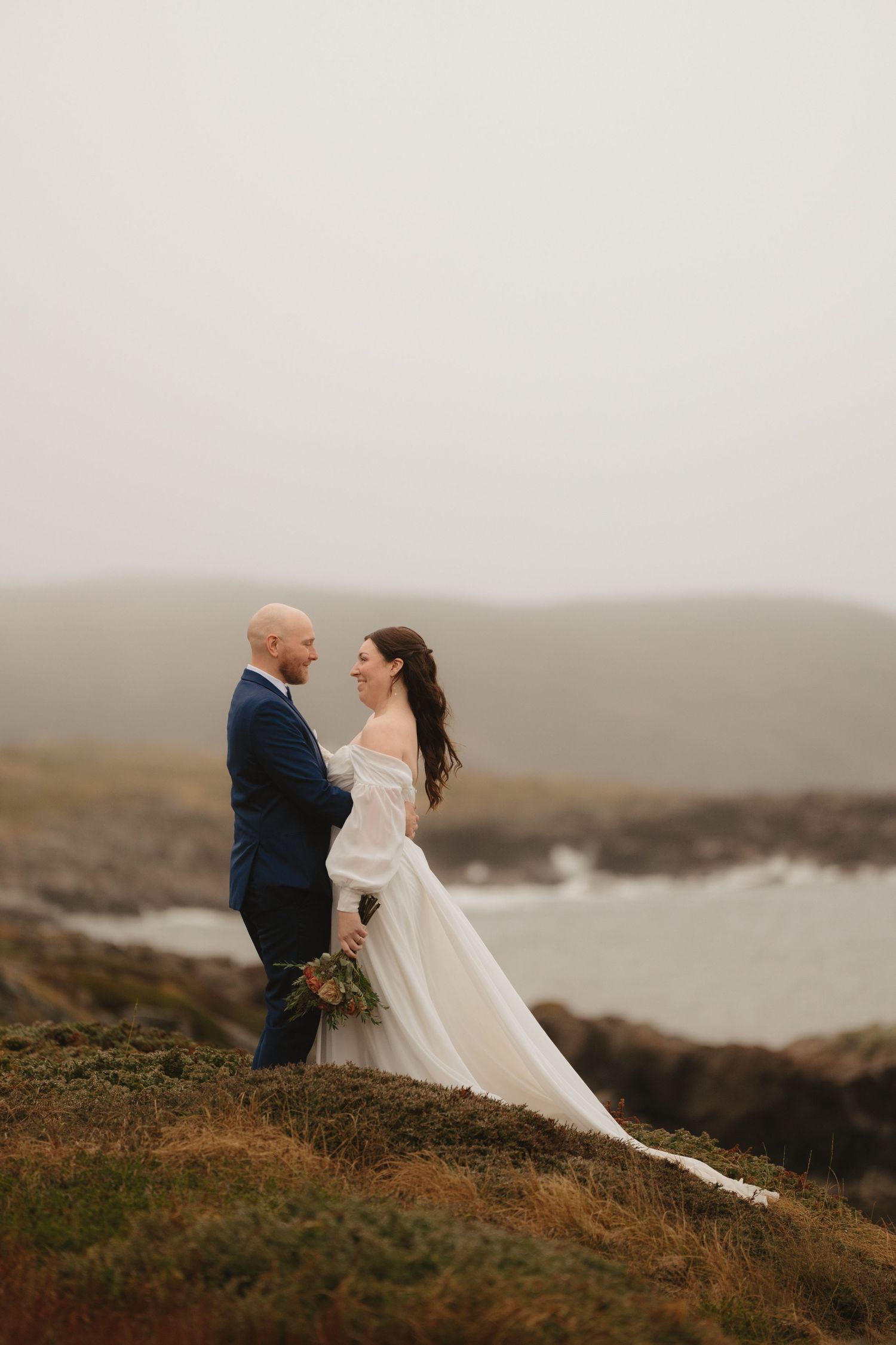 A couple in wedding attire sits together on a cliff overlooking a misty coastal landscape at sunset.