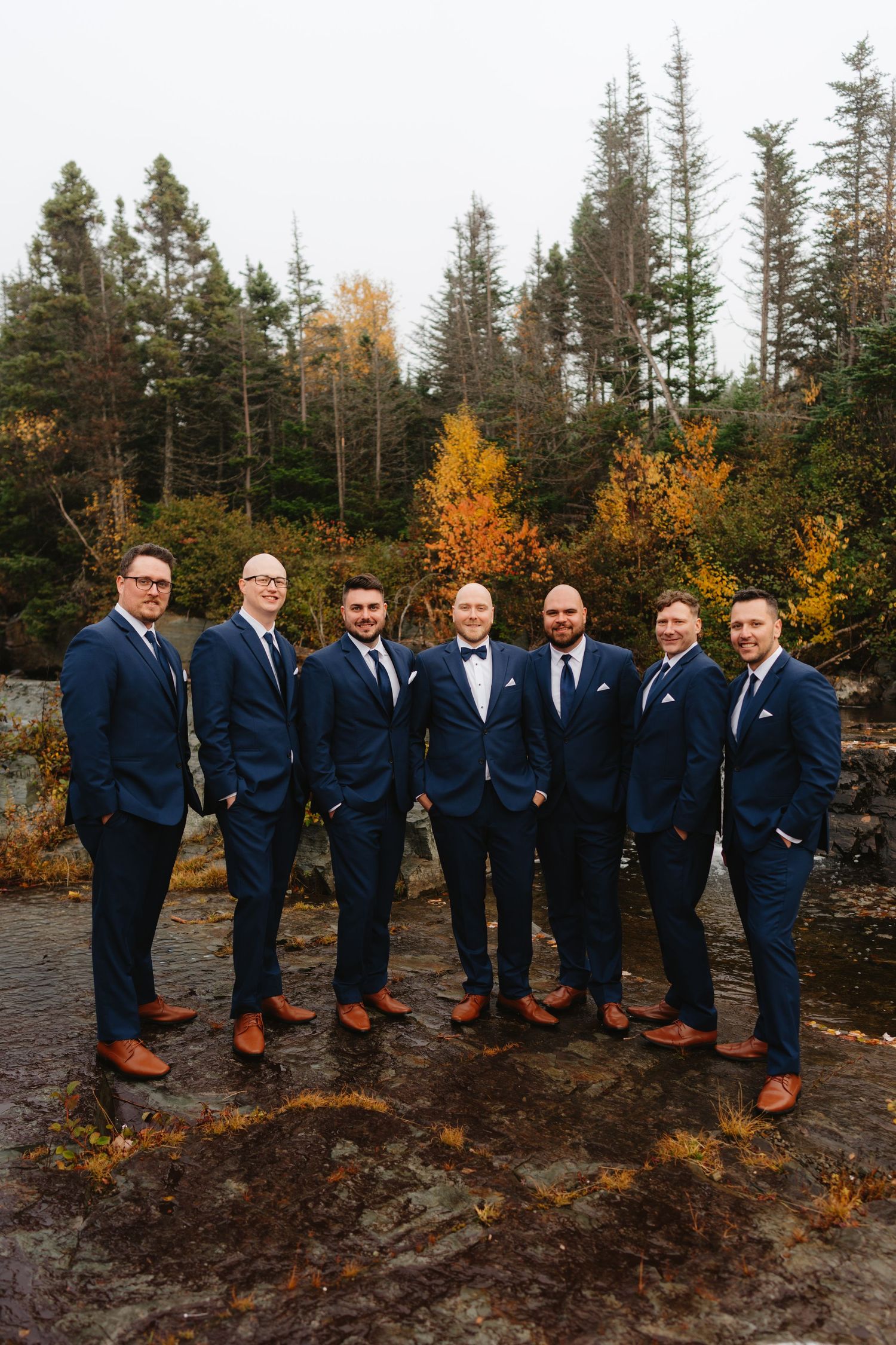 Groomsmen in navy suits and brown shoes pose together against autumn foliage and evergreen trees in outdoor setting.