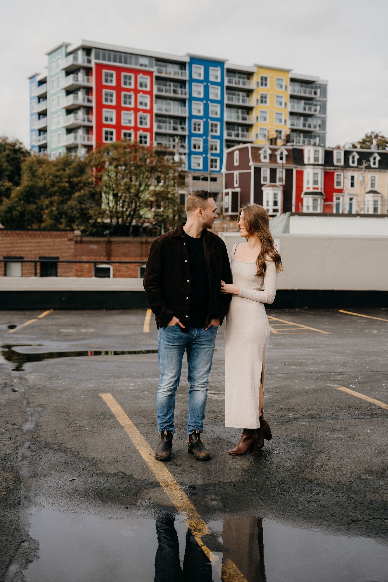 A couple shares a romantic moment in a parking lot with colorful apartment buildings in the background.