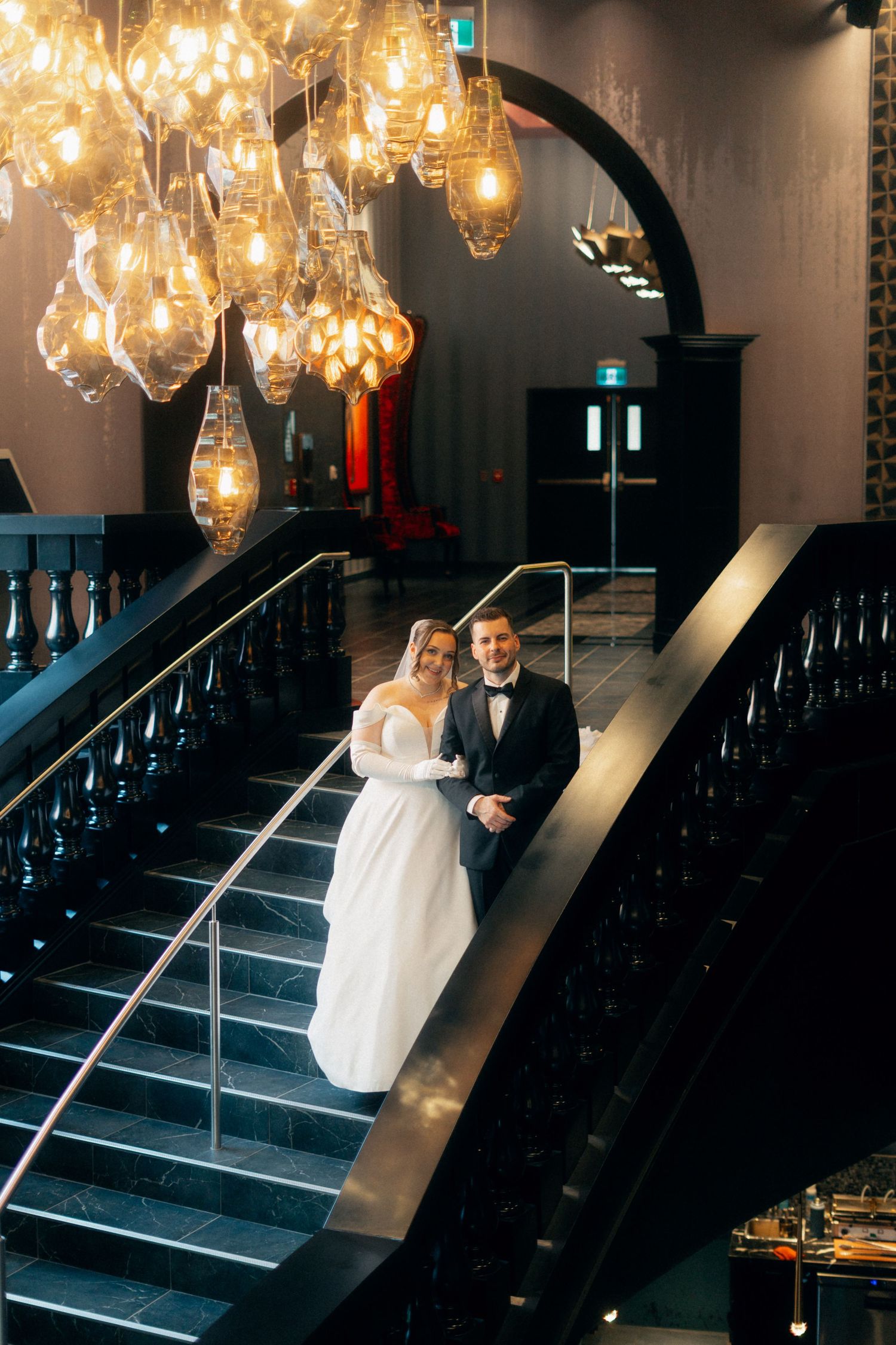 Wedding couple poses on elegant stairs under crystal chandeliers in a luxurious modern venue.