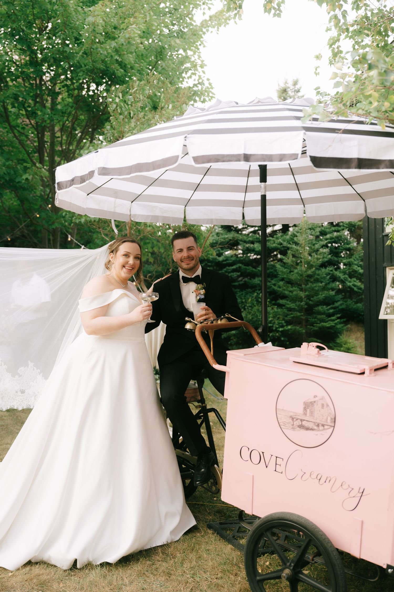 Newlyweds pose next to a pink vintage drink cart at an outdoor reception.