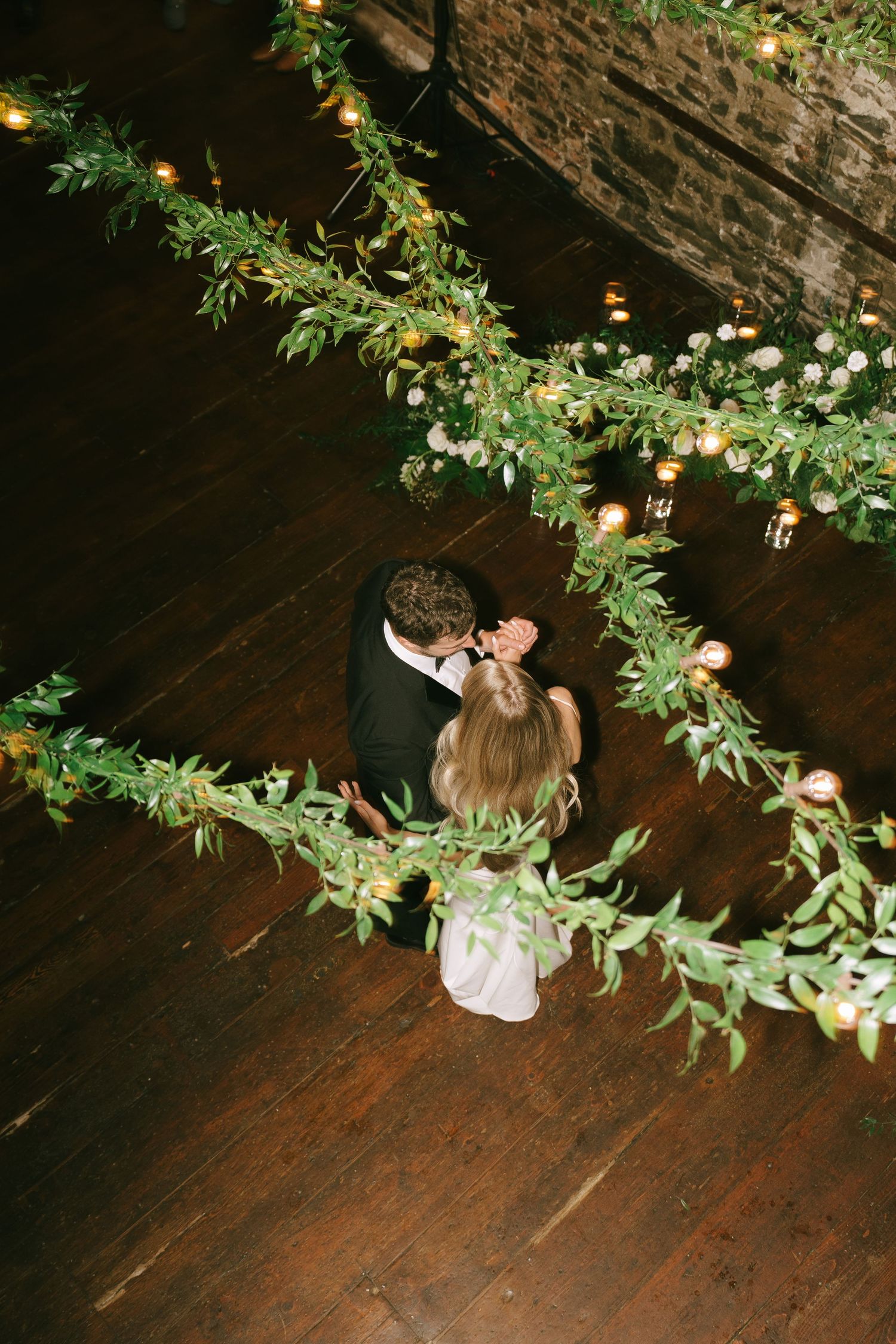 Dancing couple beneath lush green garlands and fairy lights in rustic brick venue.