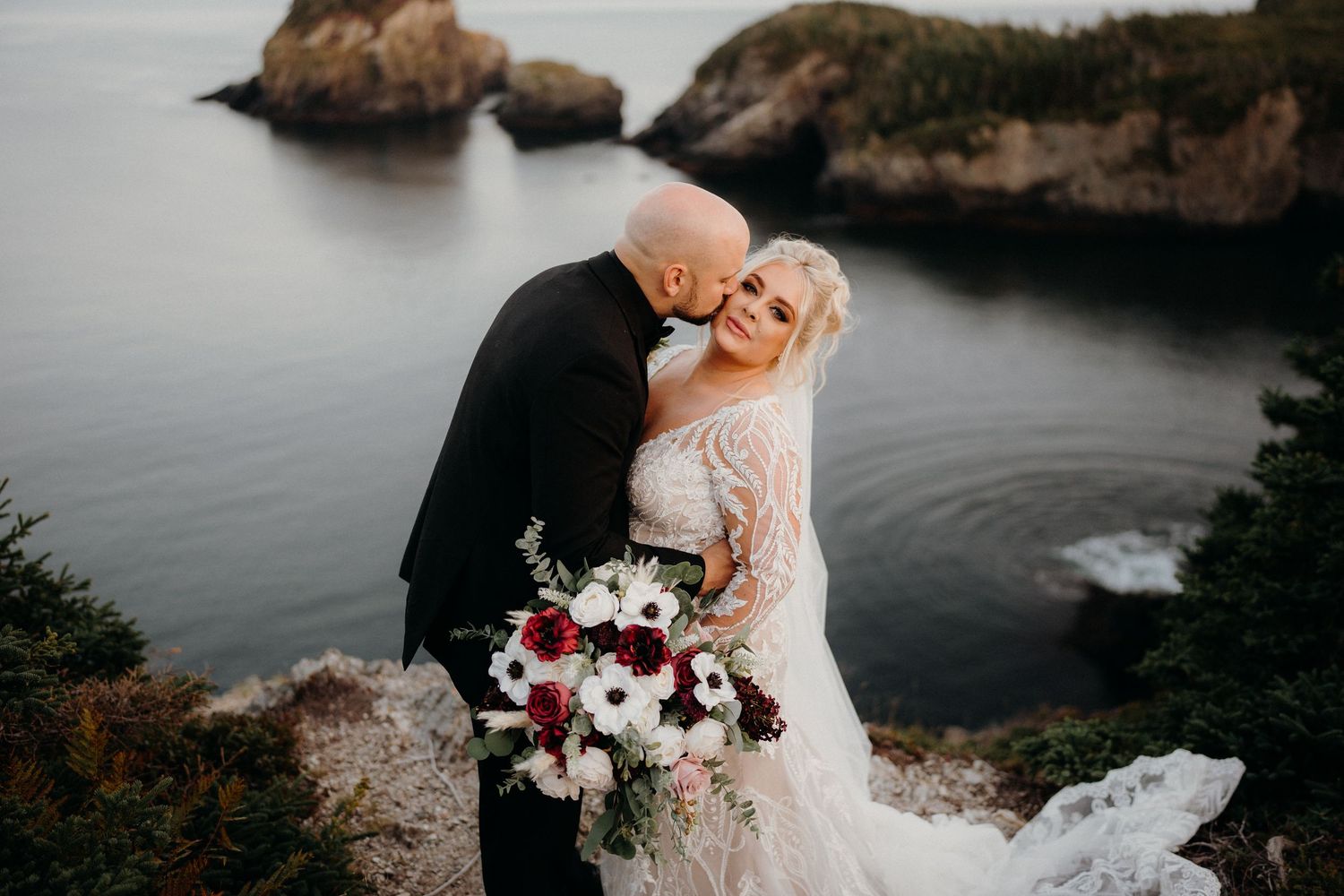 Romantic coastal wedding portrait with dramatic rocky cliffs and ocean backdrop.